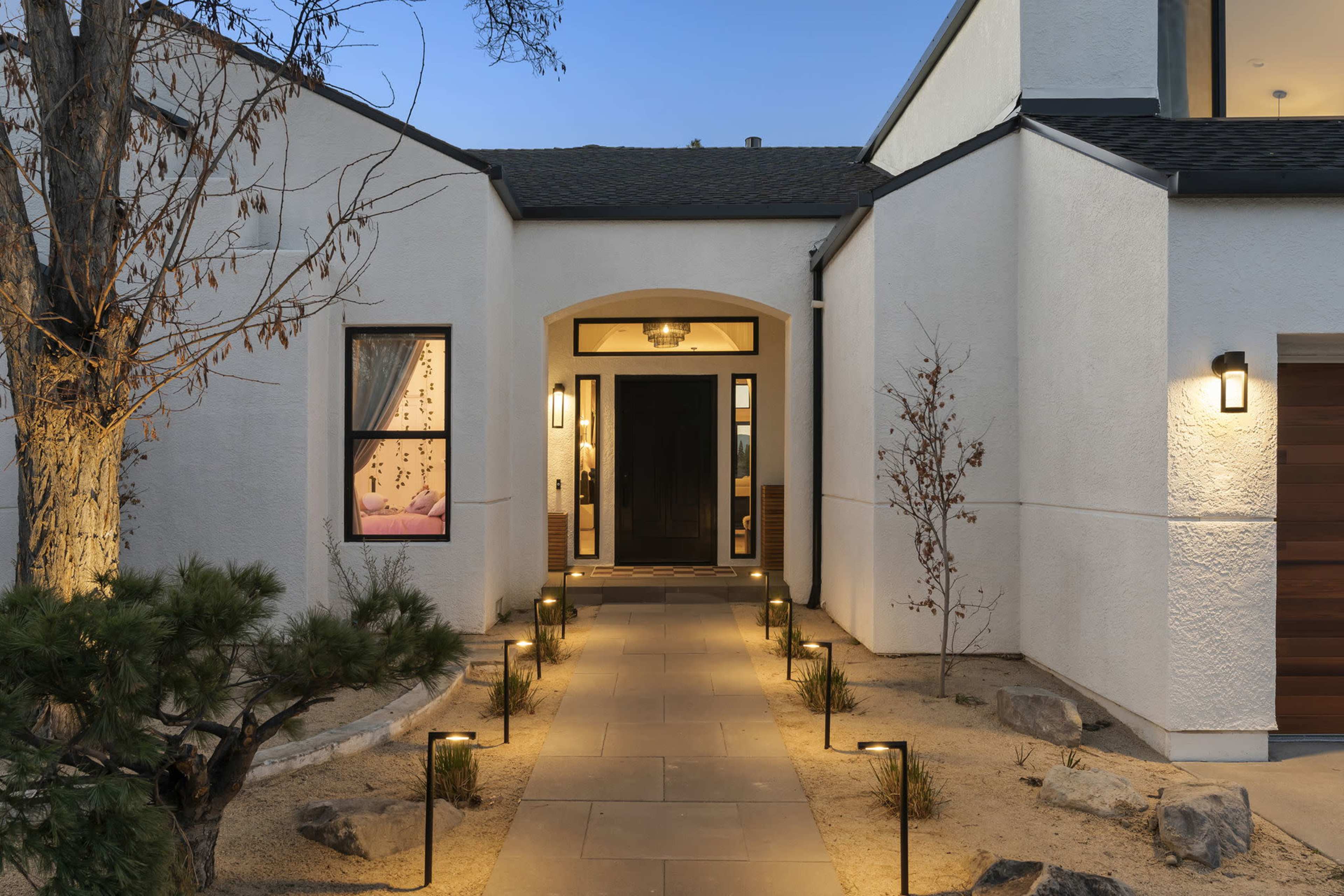 A modern house entrance with a stone path leading to a dark front door, flanked by landscaped lighting and minimal vegetation.