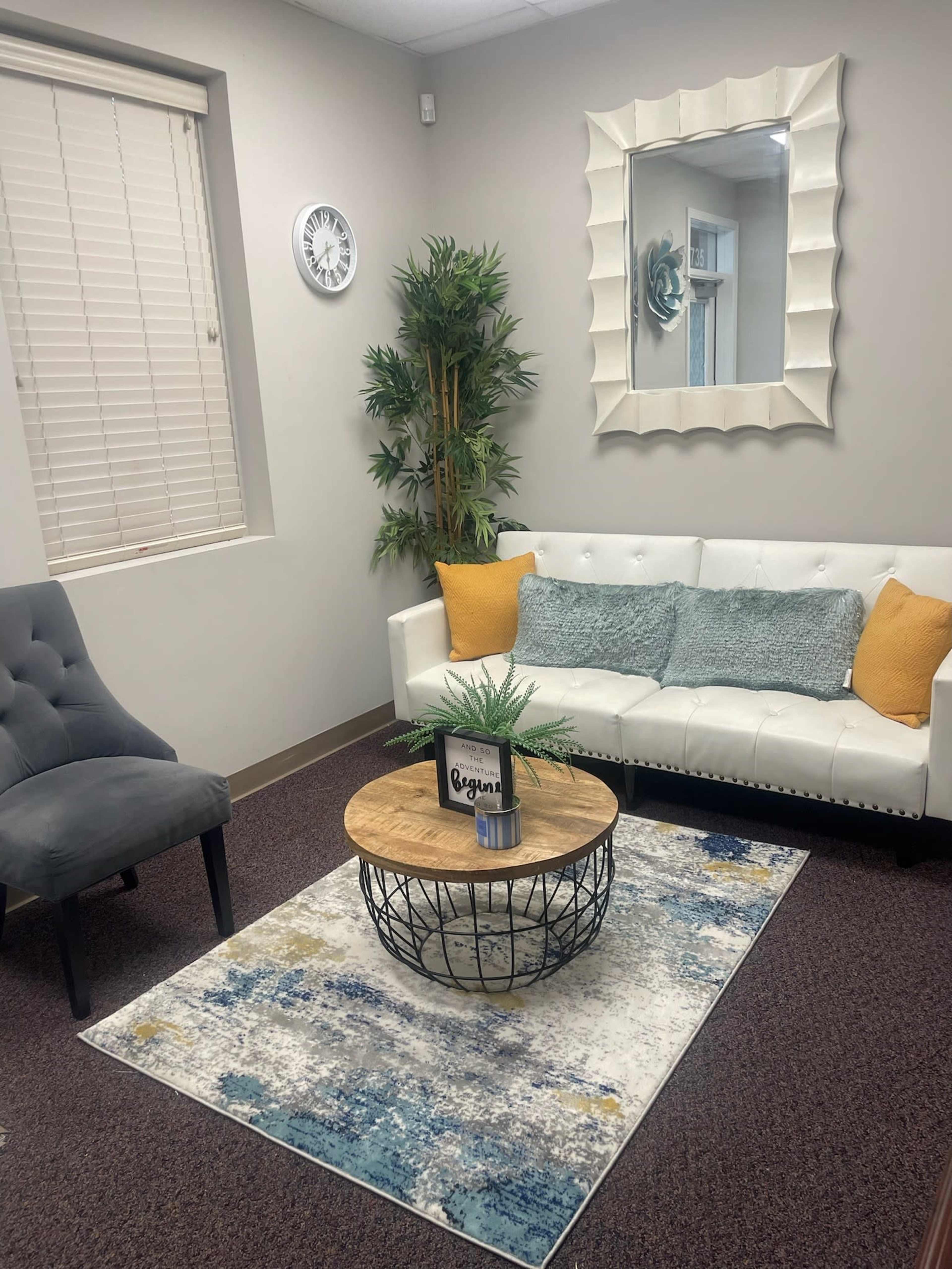A modern, stylish office waiting area with a white couch, a round wooden coffee table, a gray chair, and a large wall mirror.