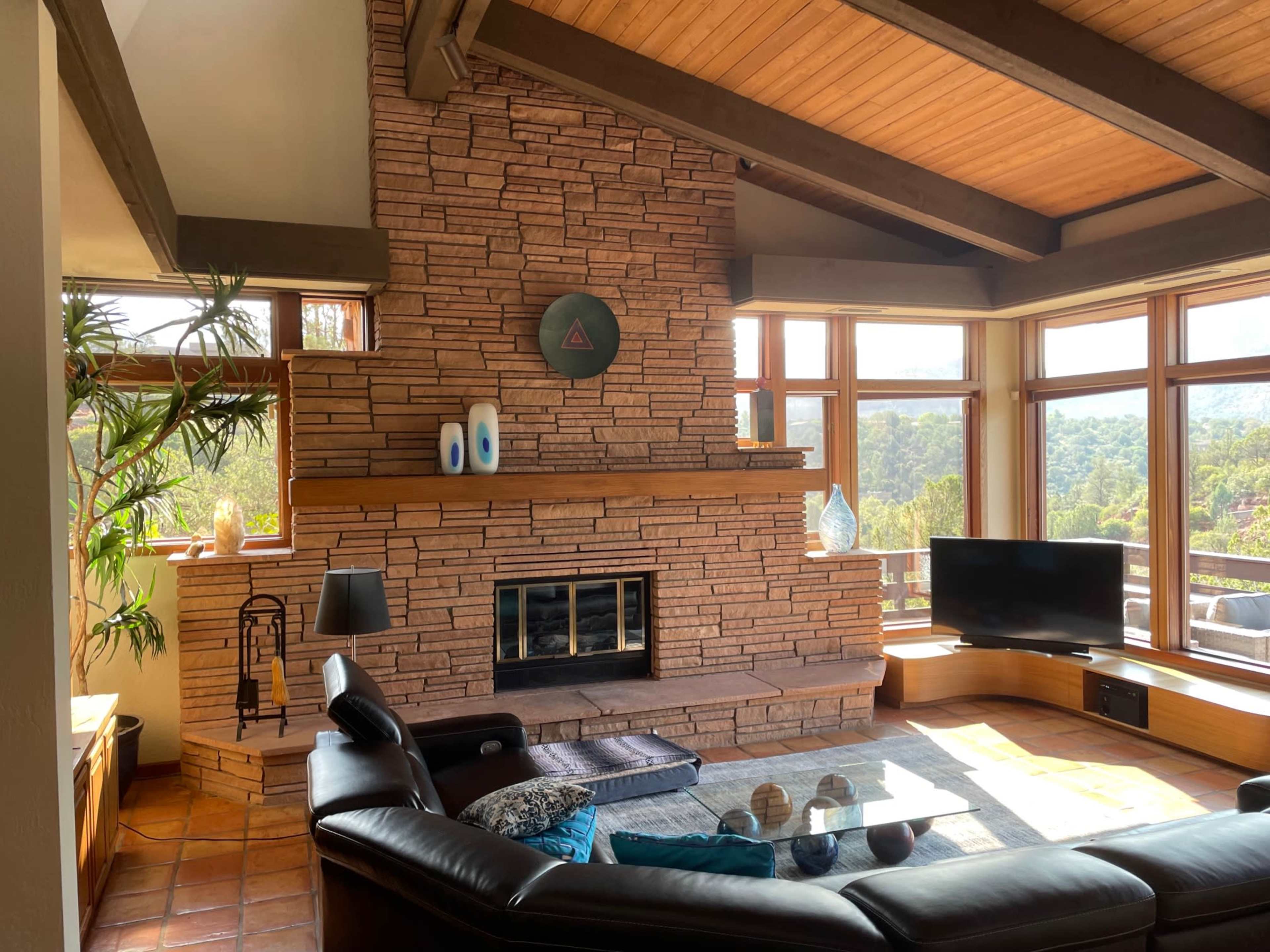 A living room with a stone fireplace, large windows offering a view of the outdoors, and a black leather sectional sofa.