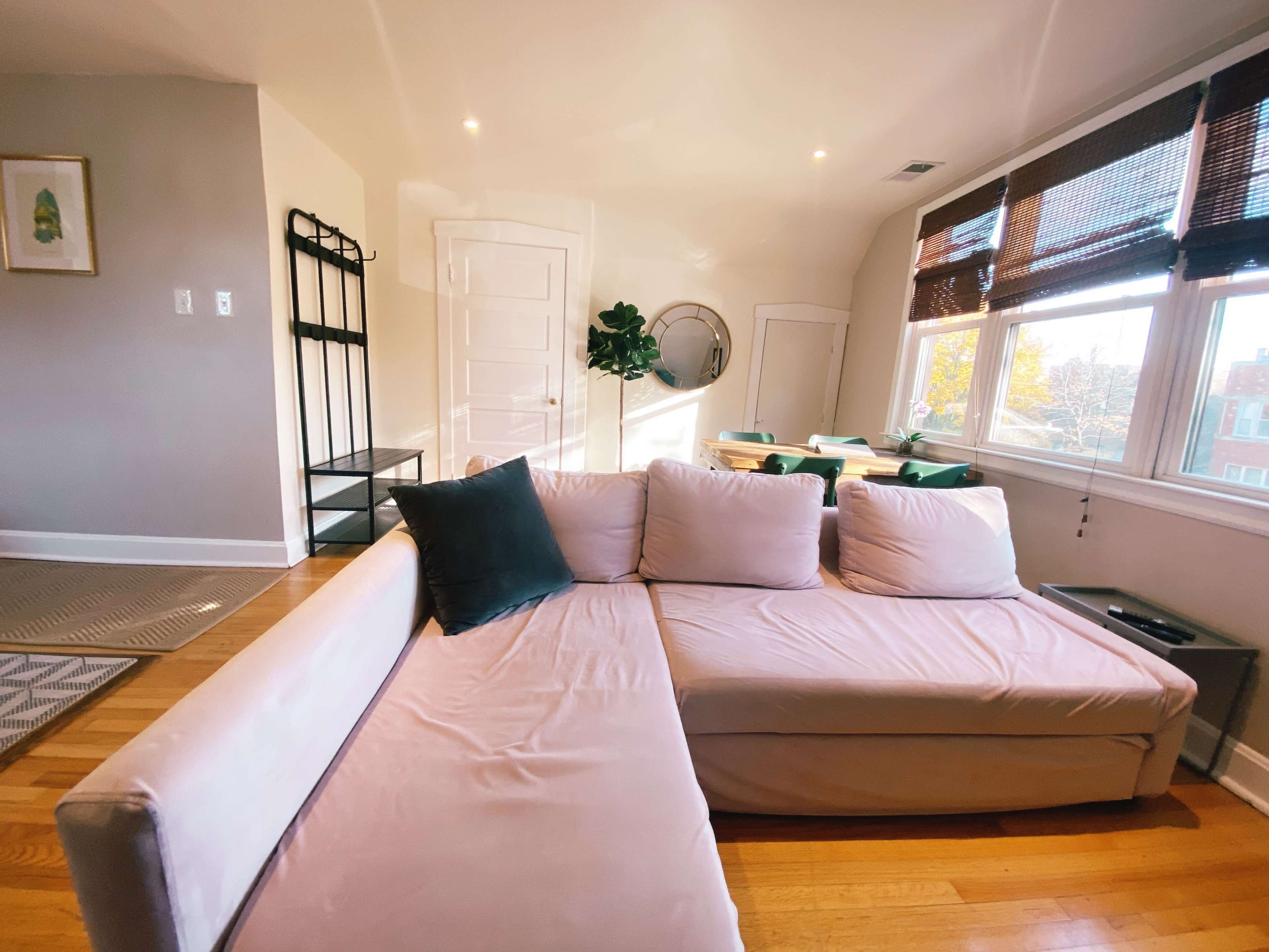 A light-colored sectional sofa is positioned in a bright room with large windows, a mirror, and a small dining area visible in the background.