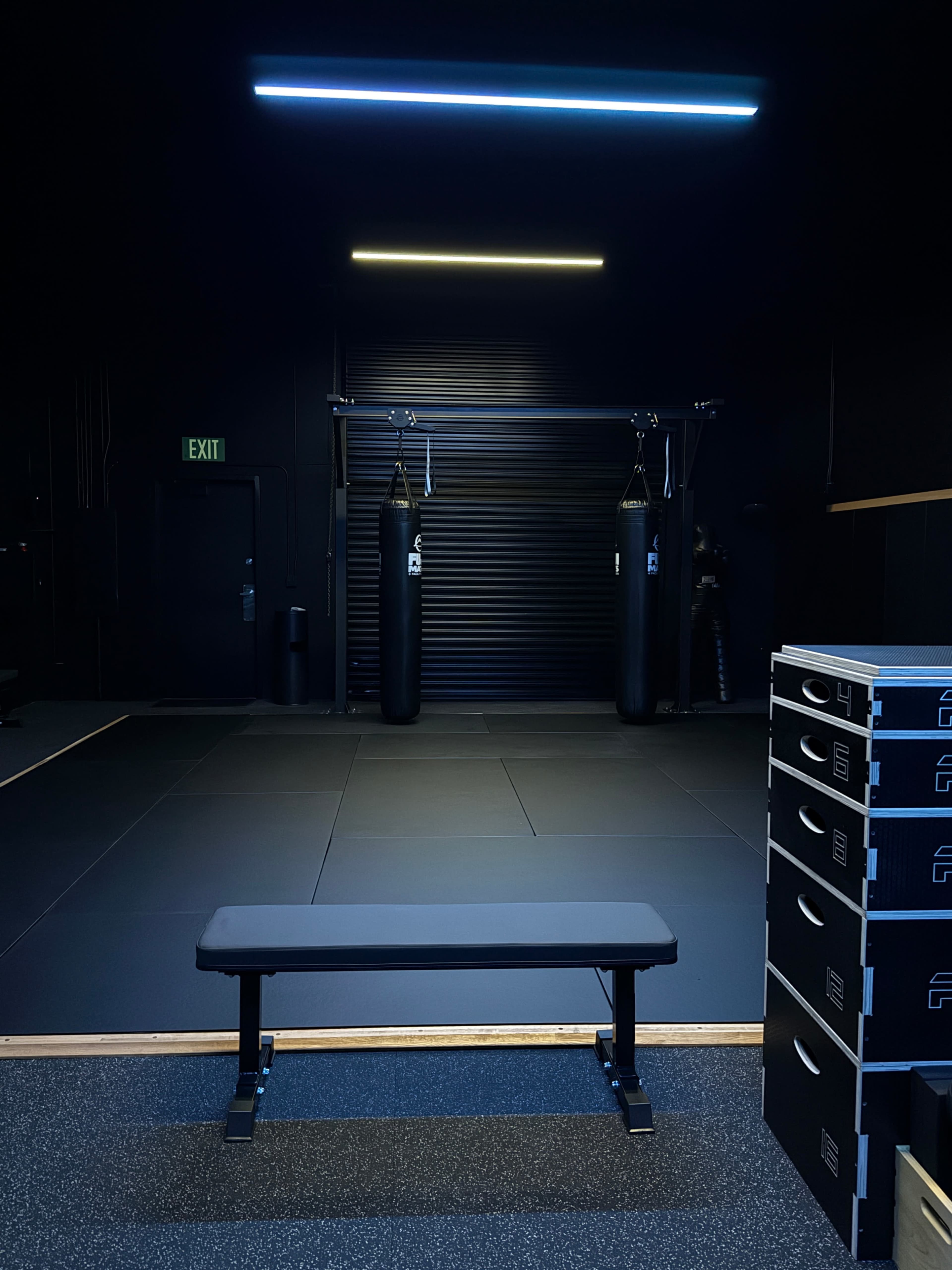 The image shows a dark gym interior featuring a bench in the foreground, two punching bags hanging in the background, and stacked boxes to the right.