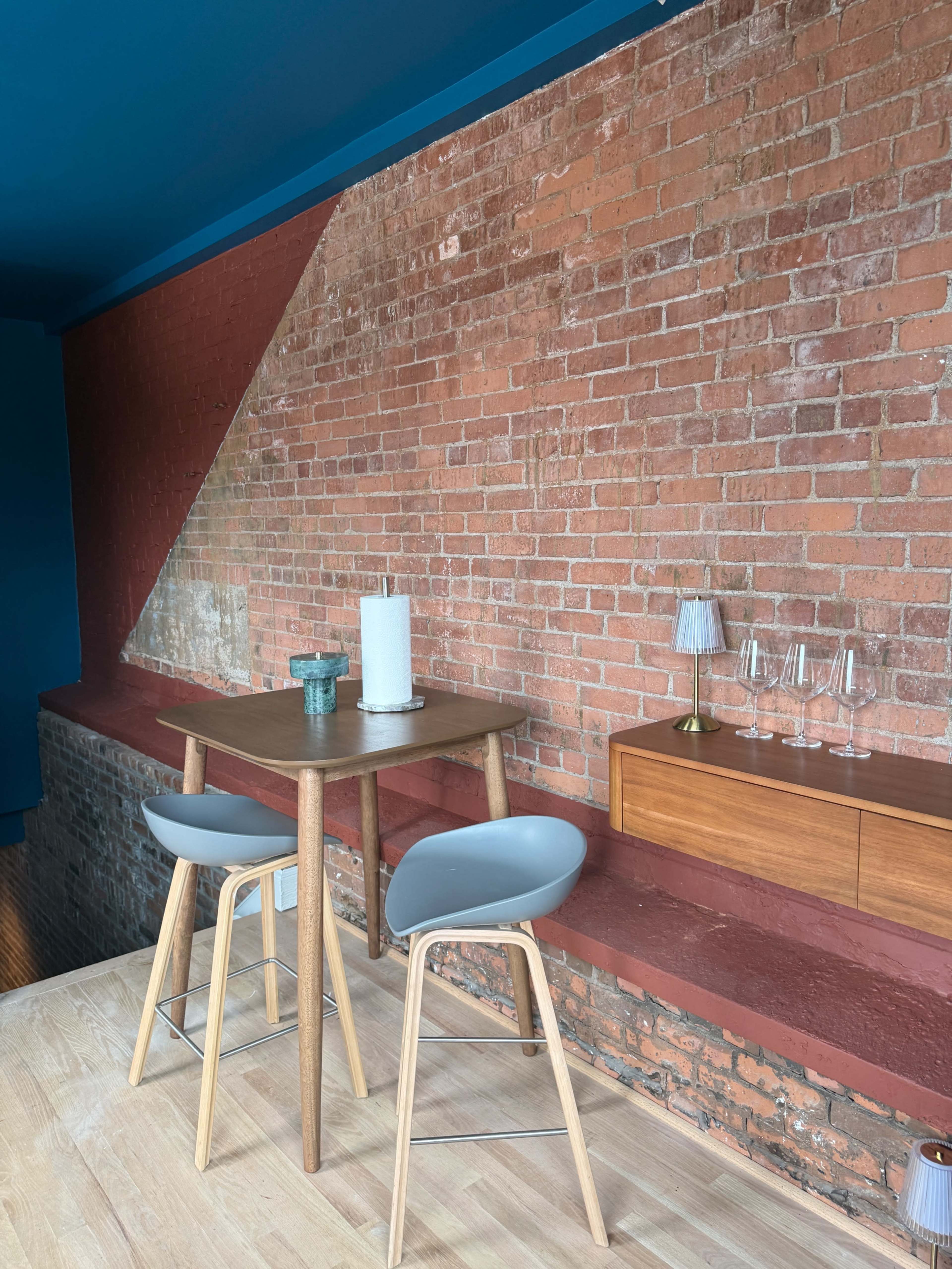 The image shows a small wooden table with two gray chairs against a backdrop of exposed brick walls in a room with blue accents.