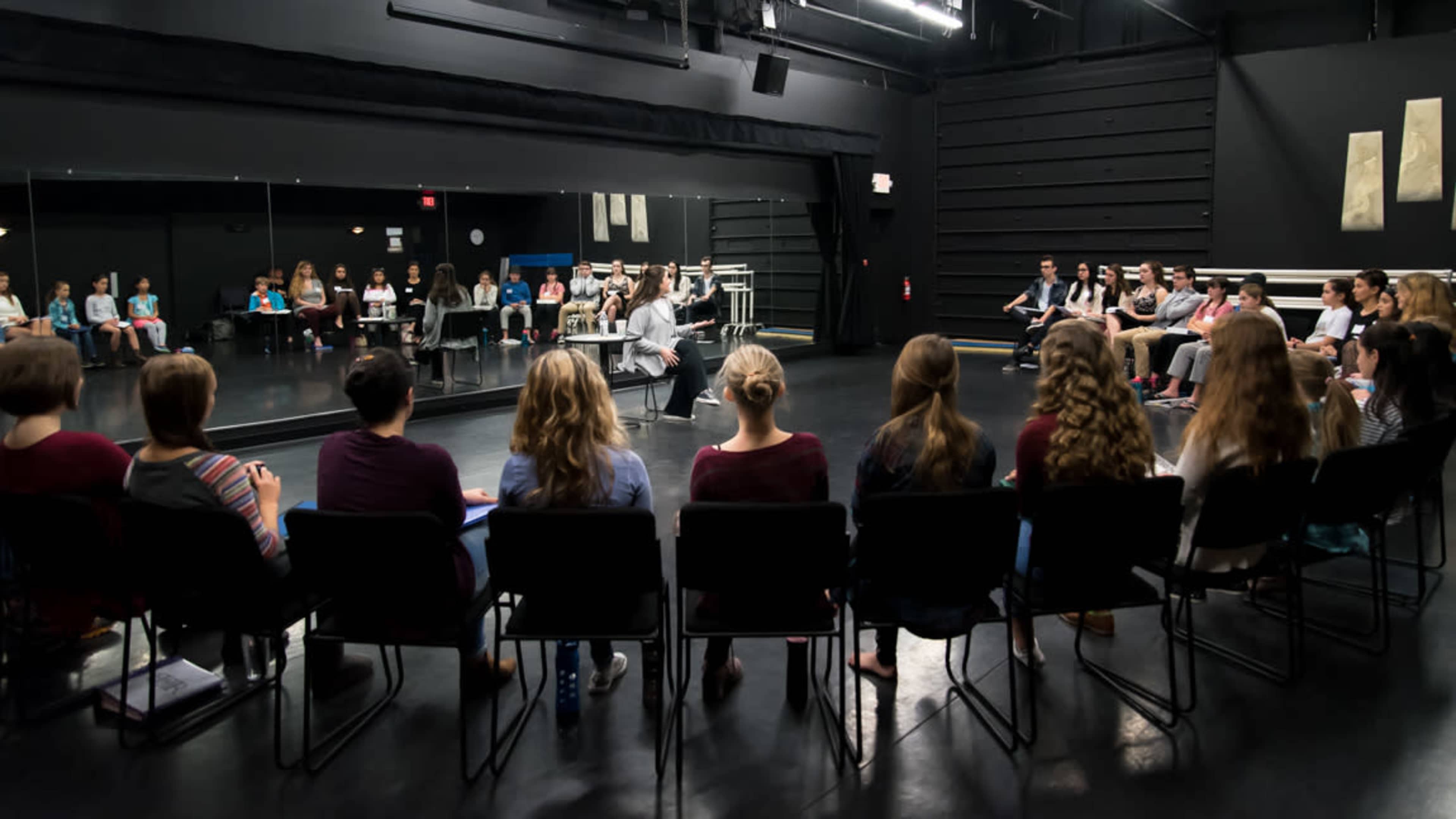 A group of students sits in chairs facing a speaker in a theater rehearsal space.