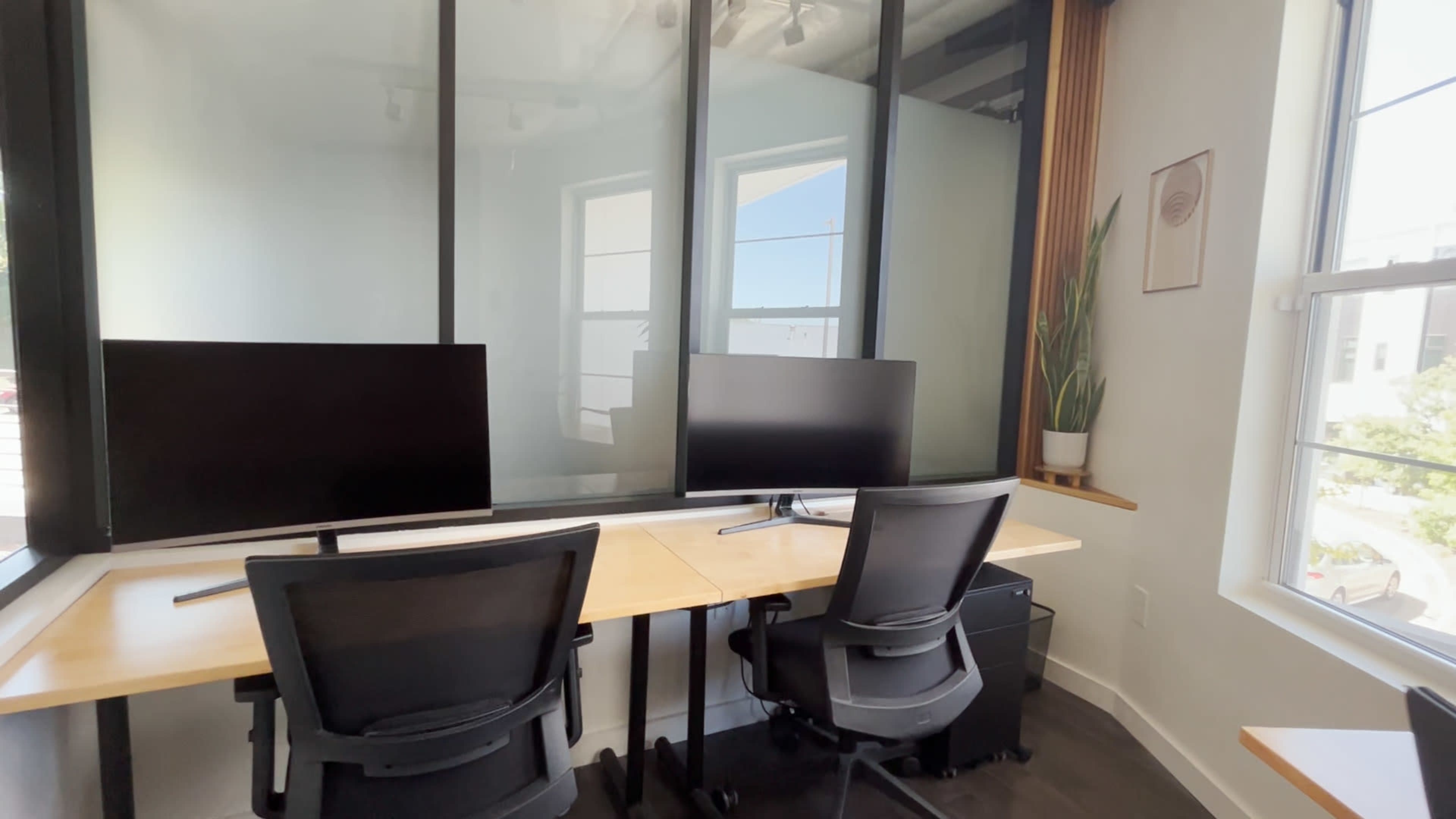 The image shows a modern office workspace with two black chairs facing two computer monitors on a wooden desk, surrounded by glass walls and a potted plant near a window.