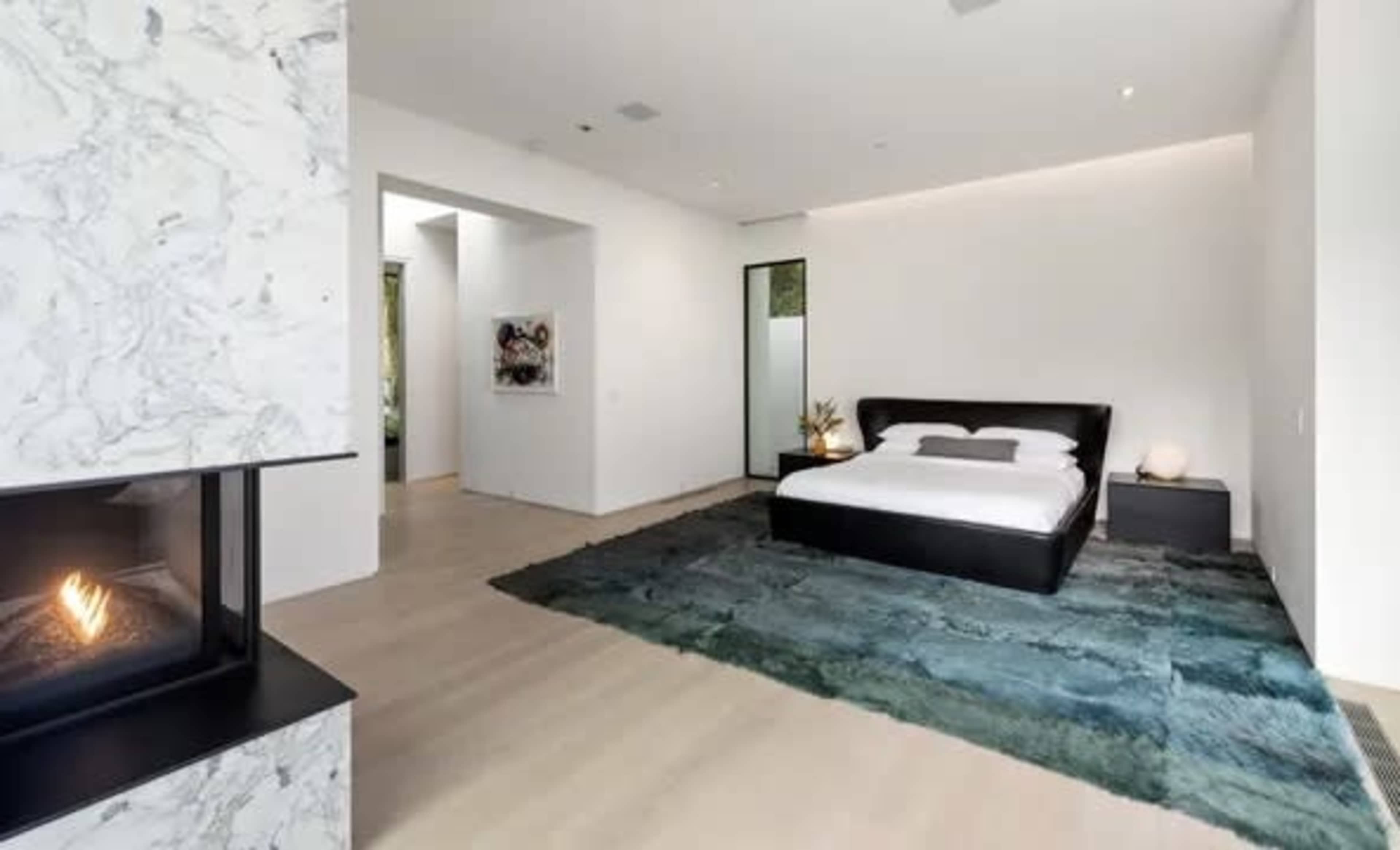 A modern bedroom featuring a black bed on a textured rug, a minimalist fireplace, and white walls with a doorway leading to another room.