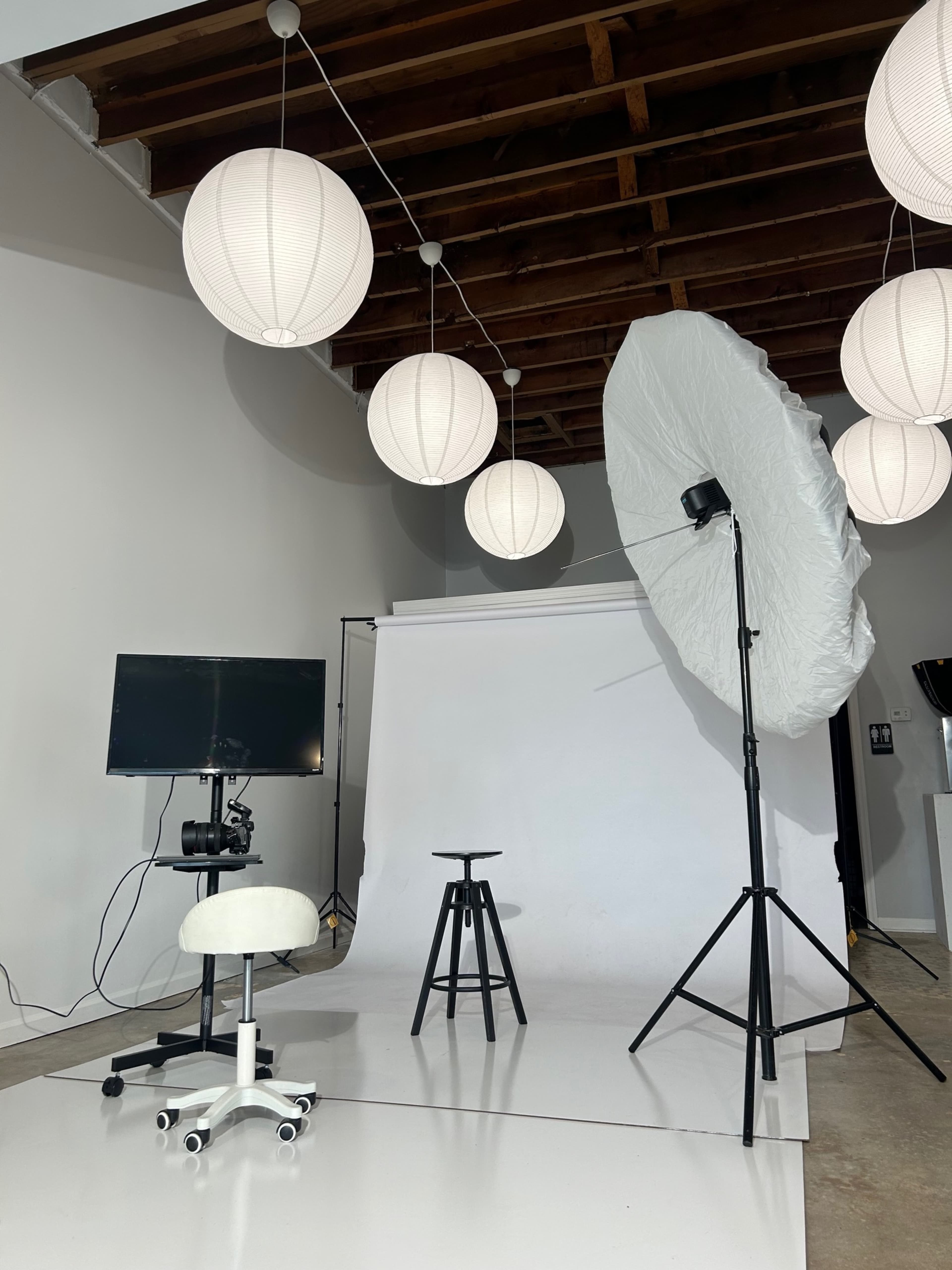 The image shows a photography studio setup featuring a white backdrop, two stools, a softbox light, and decorative paper lanterns hanging from the ceiling.