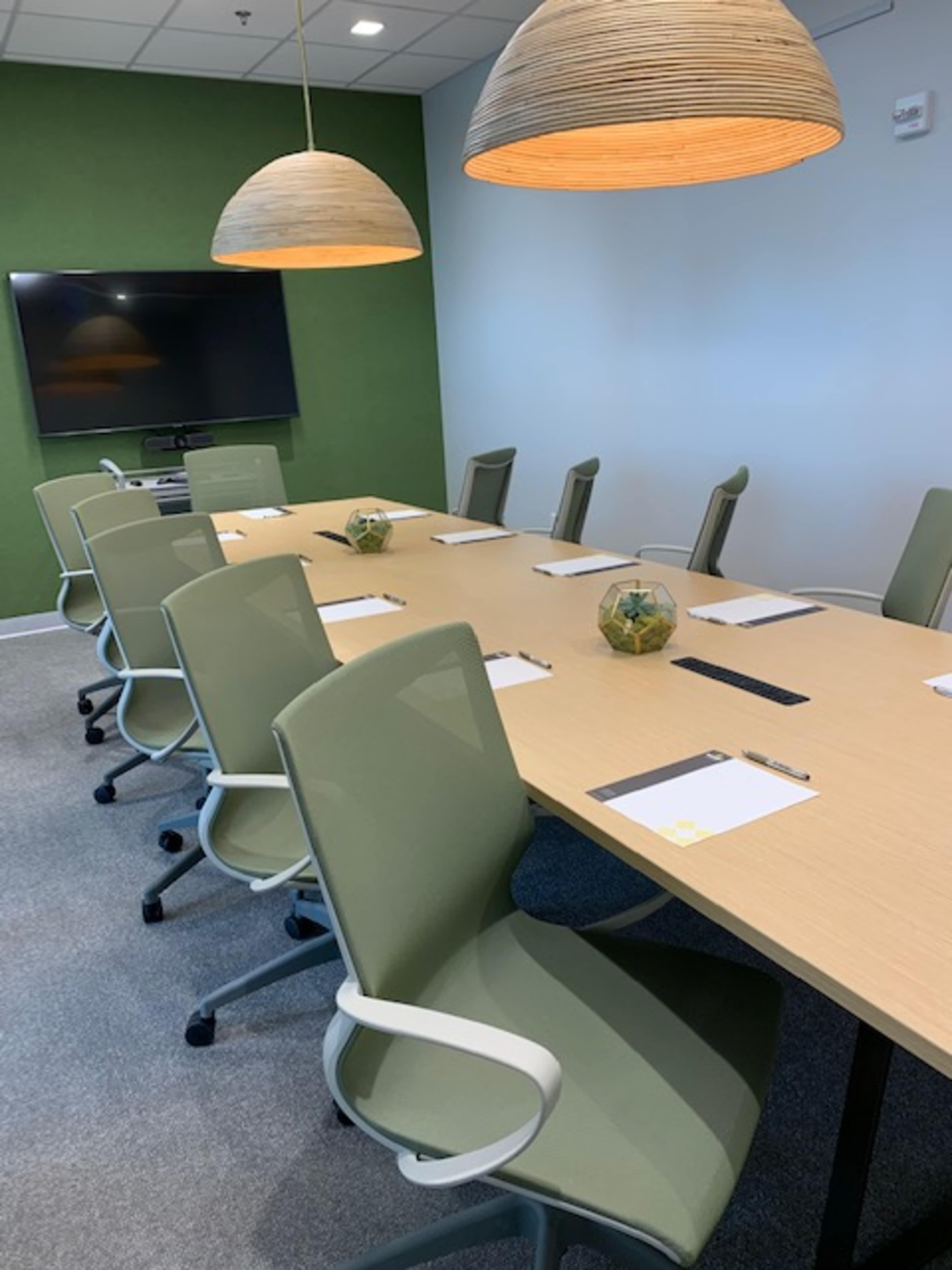 A conference room features a long wooden table surrounded by green chairs, with two pendant lights overhead and a television on the wall.