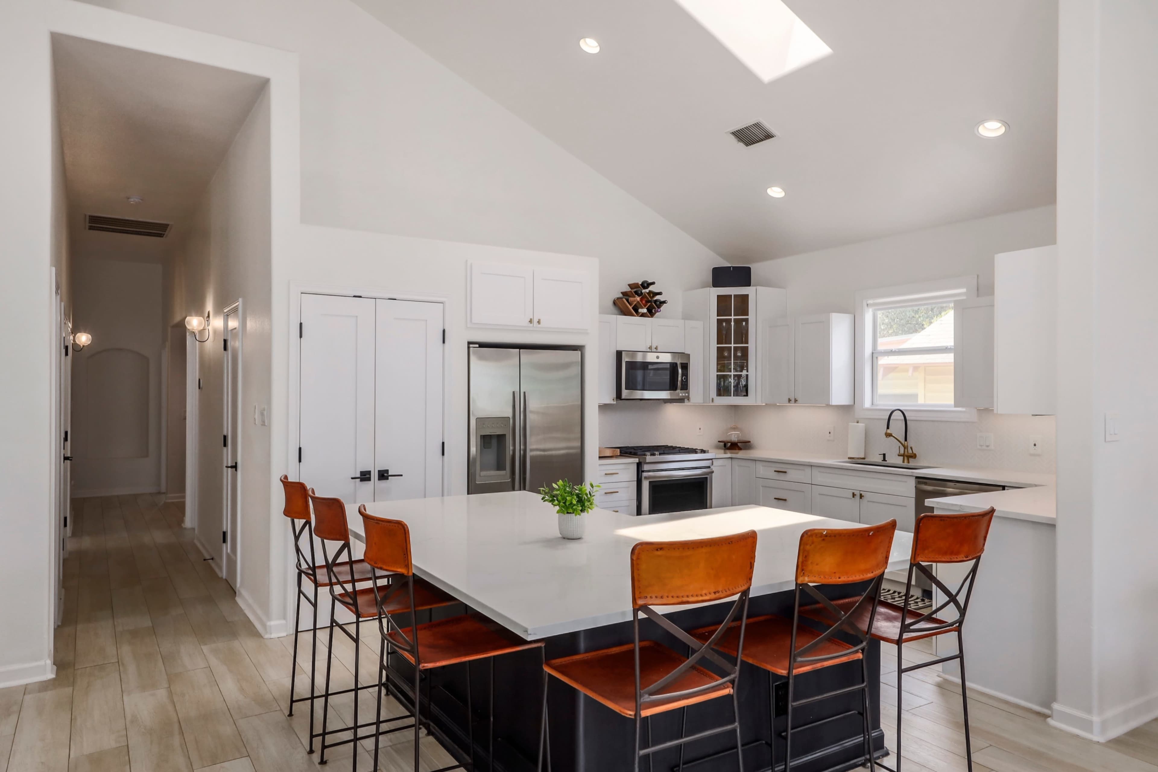 The image shows a modern kitchen with a large island, eight metal stools, stainless steel appliances, and white cabinets under a high ceiling.