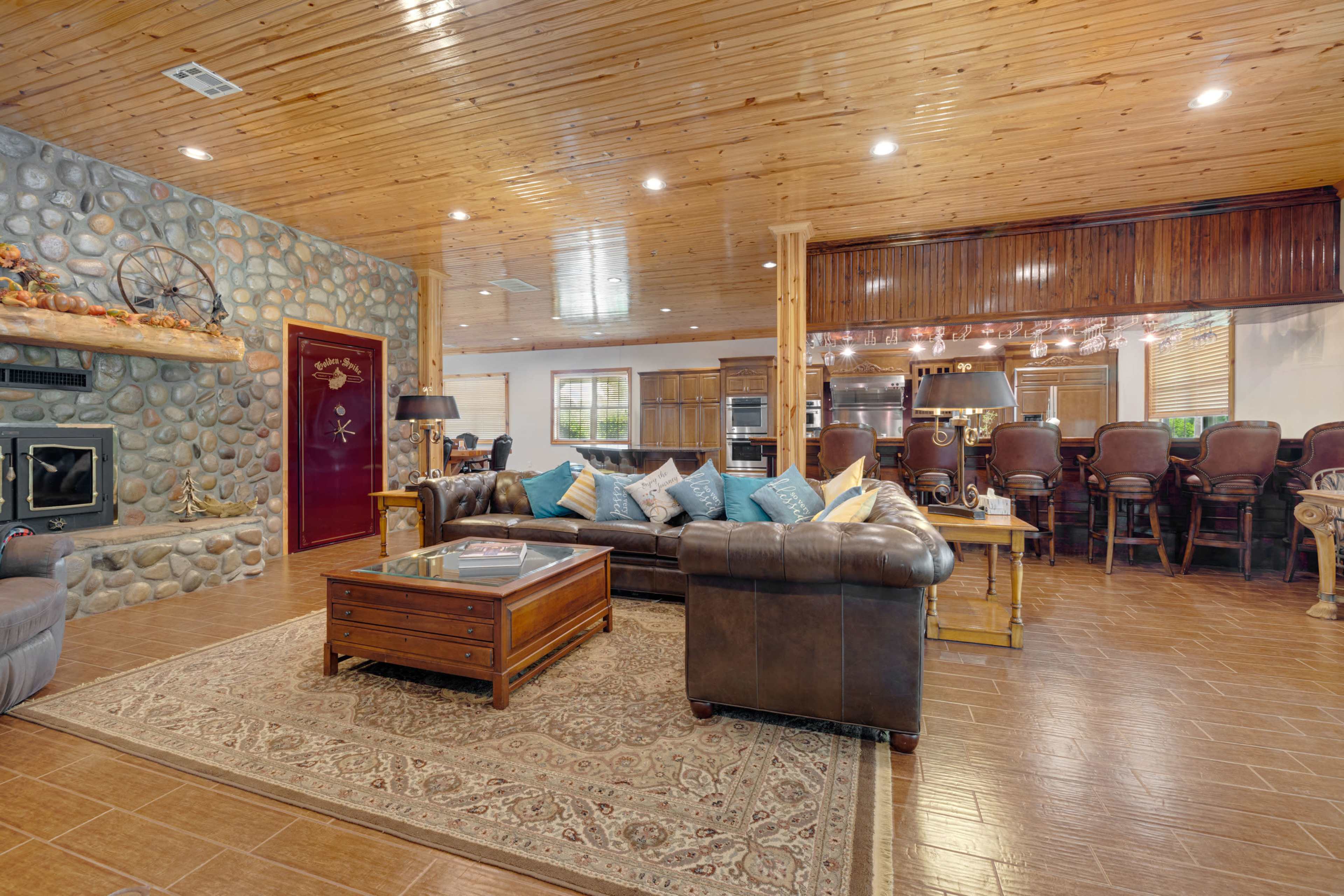 A spacious living room featuring a stone fireplace, leather sofas with decorative pillows, and a dining area with bar stools in the background.