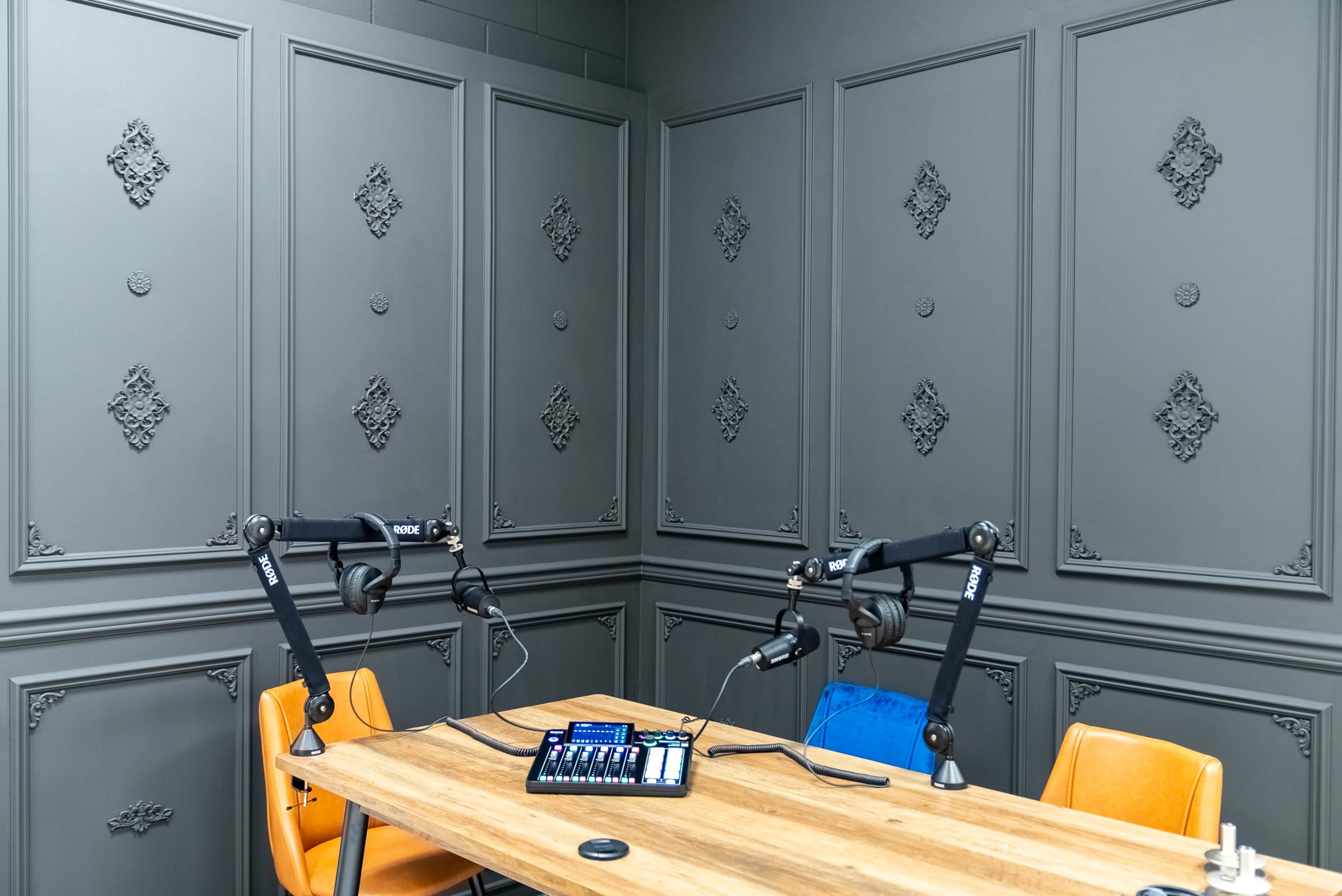 A small recording studio features a wooden table with two microphone stands and audio equipment against a textured gray wall.