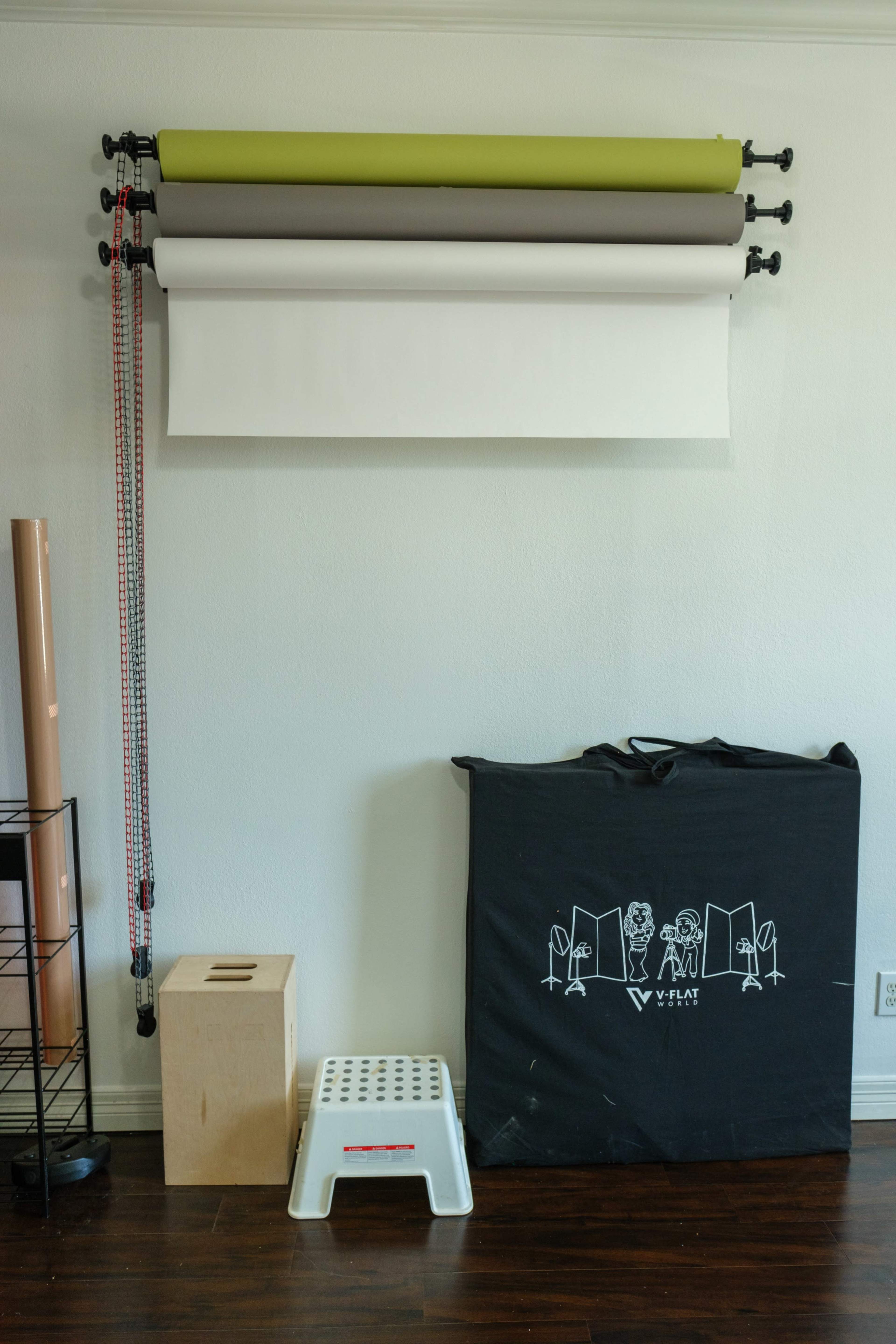 A set of paper backdrops in various colors is mounted on a wall, with a small storage box and a step stool on the floor beneath them.