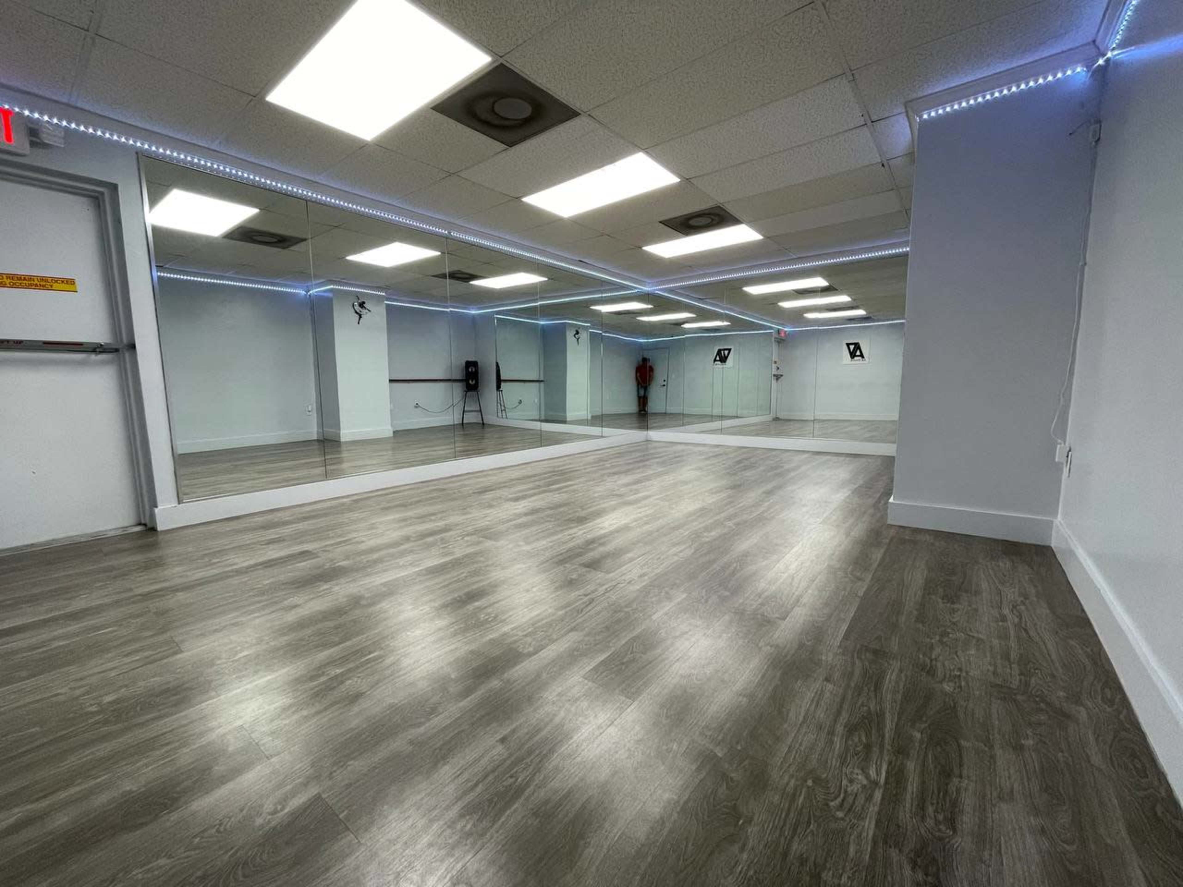 A spacious dance studio with mirrored walls and wooden flooring, equipped with overhead lighting.
