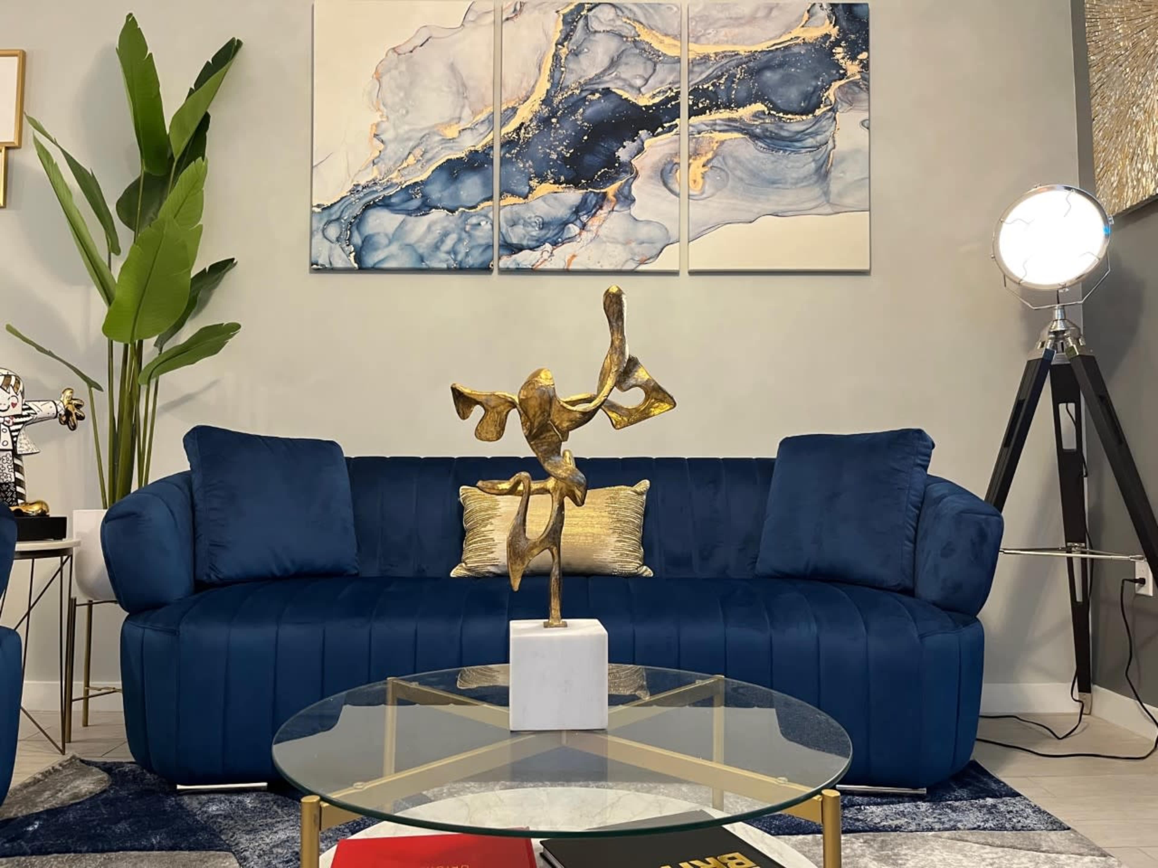 The image features a modern living room with a blue couch, a glass coffee table displaying books, and a decorative sculpture on a pedestal, complemented by abstract artwork on the wall.