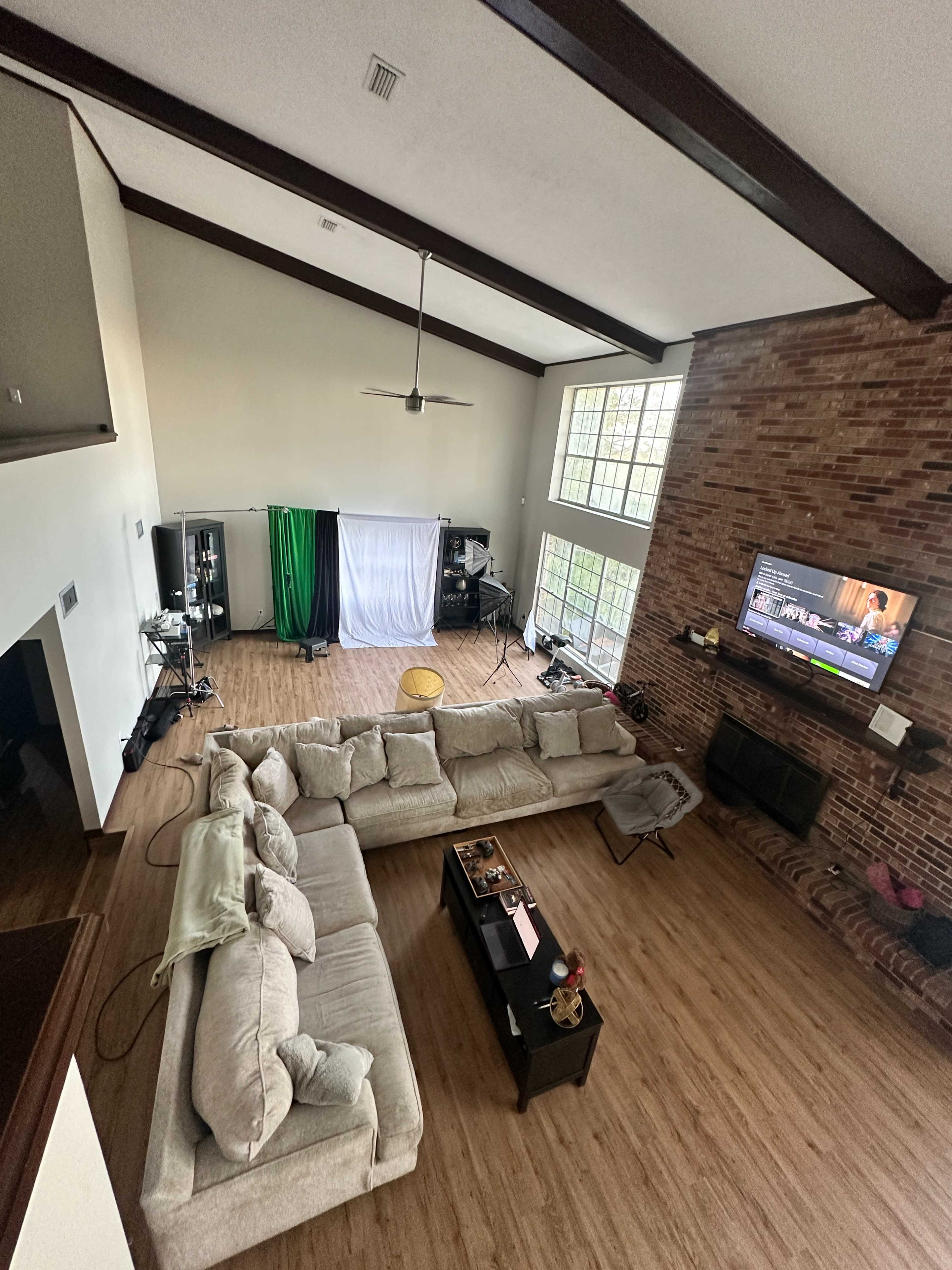 The image shows a spacious living room with high ceilings, featuring a large sectional sofa, a coffee table, a fireplace, and equipment for filming in one corner.