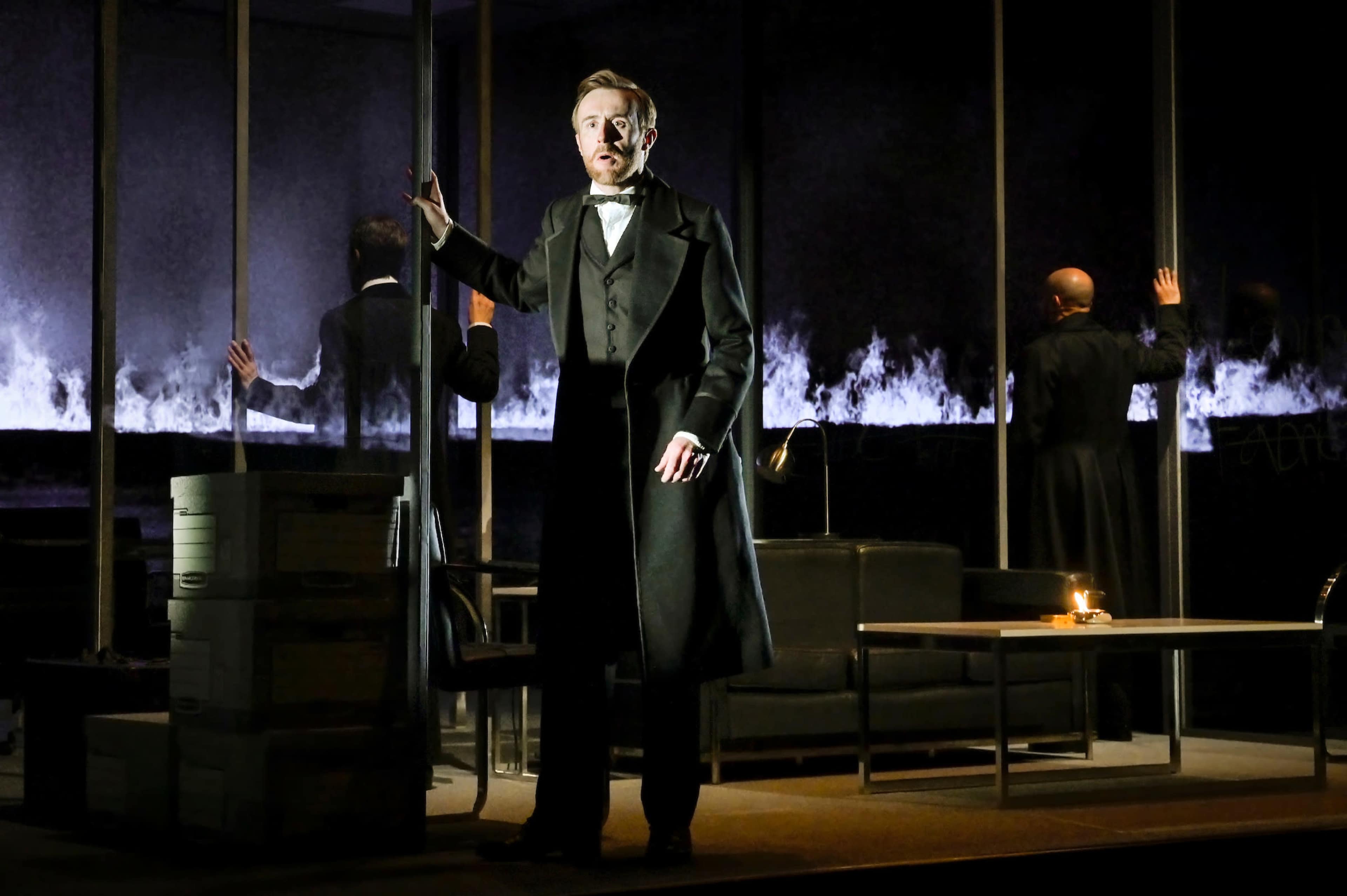 A man in formal evening attire stands in a dimly lit room, with other figures faintly visible in the background, surrounded by stacks of boxes and a candle on a table.