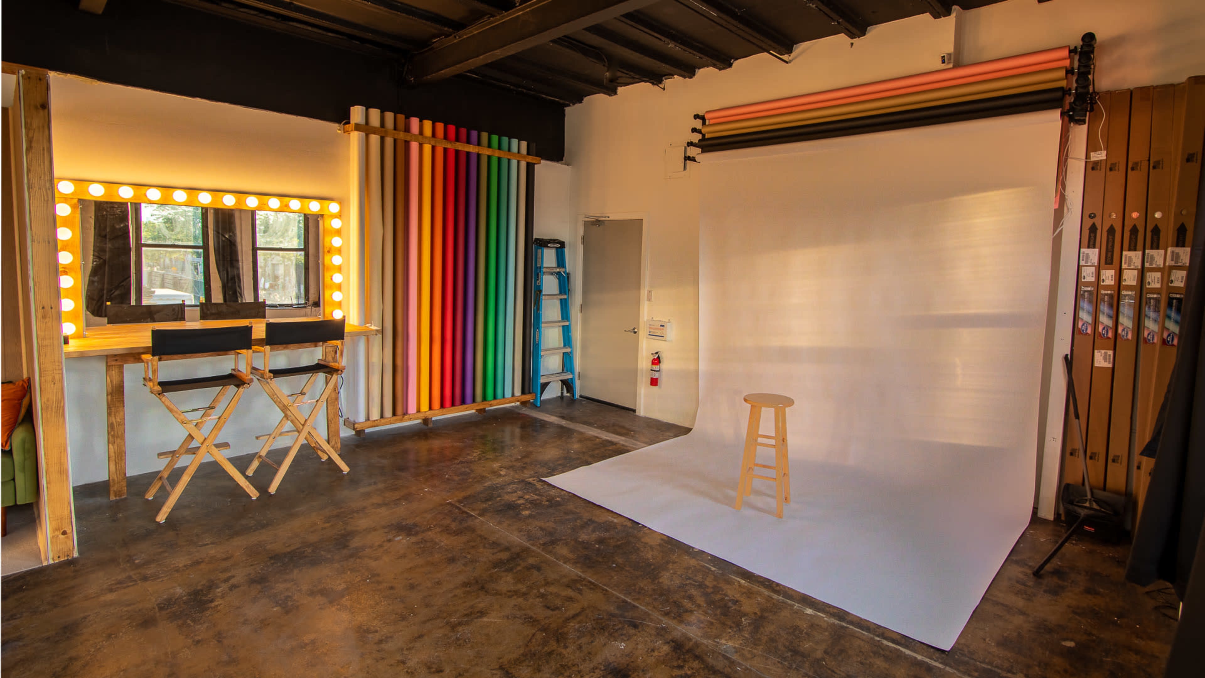 A photography studio with a backdrop setup, a wooden stool on a white floor, and colorful rolls of paper hanging on the wall next to a lighted vanity mirror.