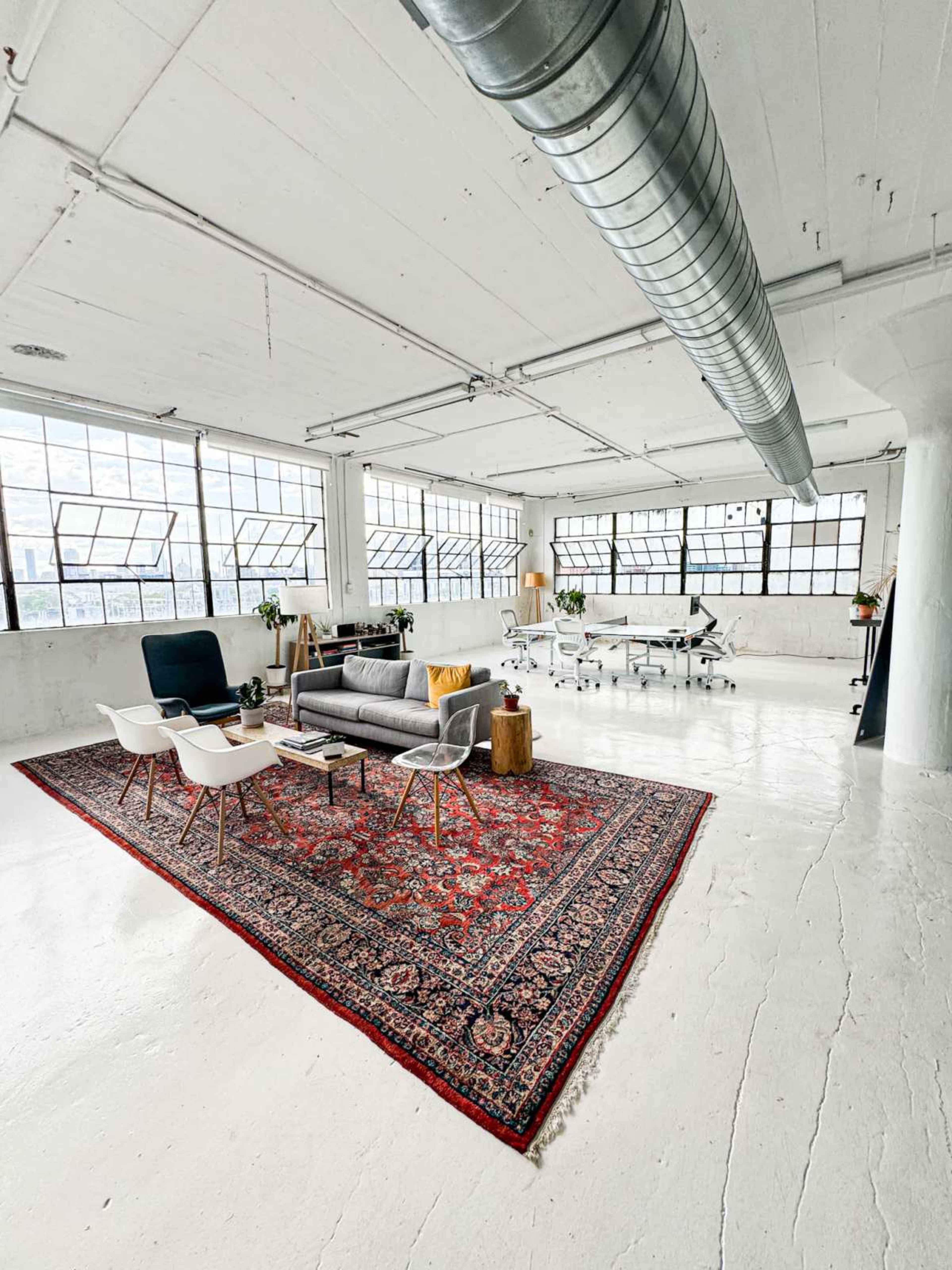 A bright, open office space features a gray sofa and white chairs on a red patterned rug, surrounded by white walls and large windows.