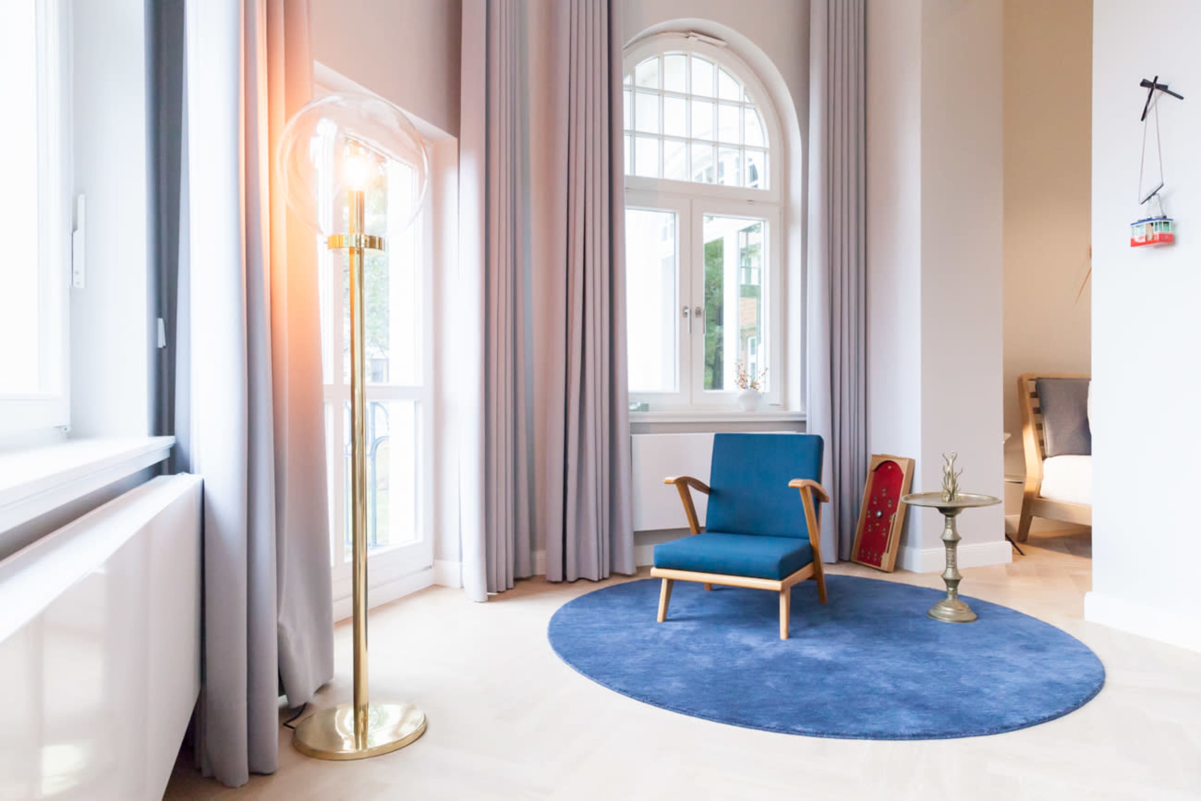 A modern room featuring a blue chair, a round blue rug, a metallic side table, and large windows with sheer curtains.