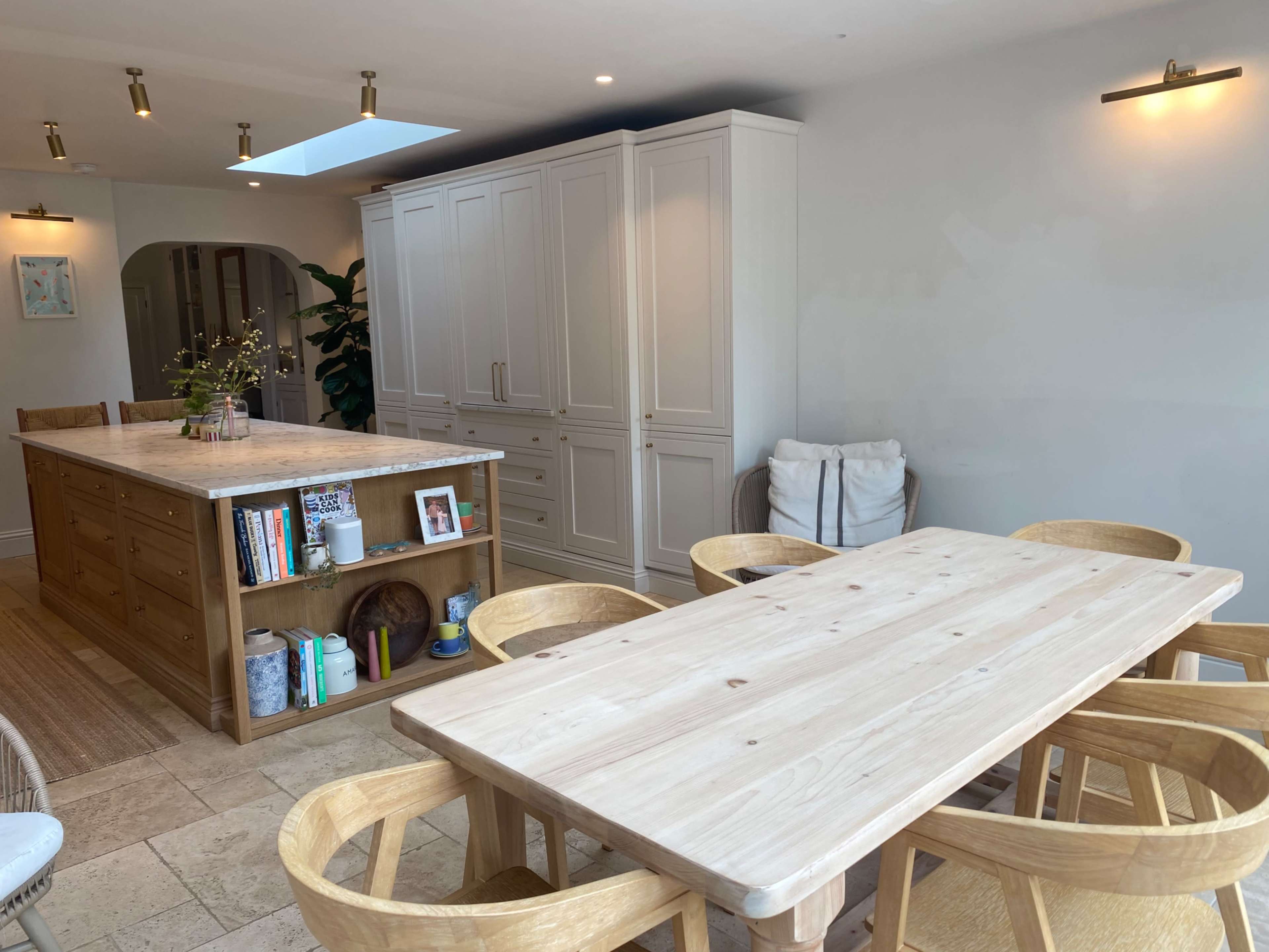 The image shows a modern kitchen with a large wooden dining table surrounded by six chairs, a central island with a marble countertop, and cabinetry along the walls.
