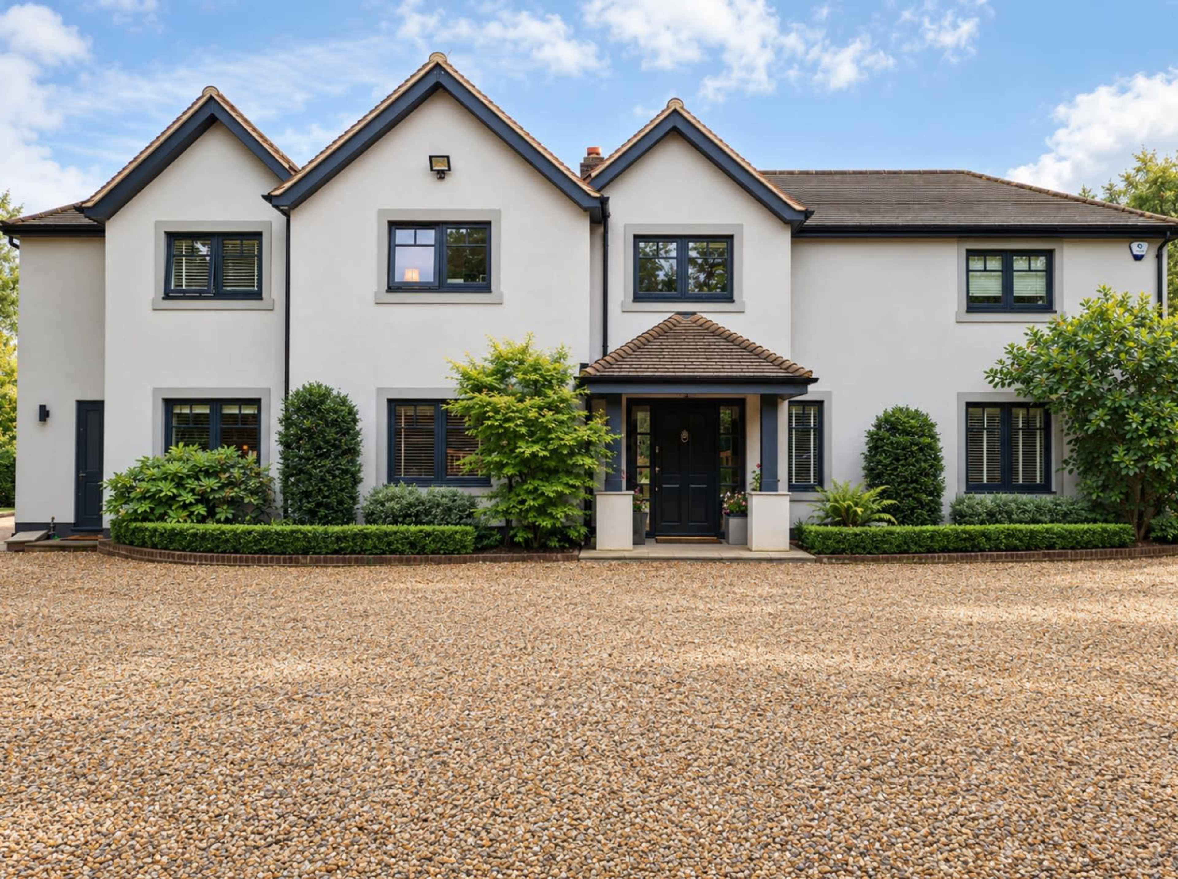 The image shows a large, modern two-story house with a gravel driveway and landscaped greenery surrounding it.