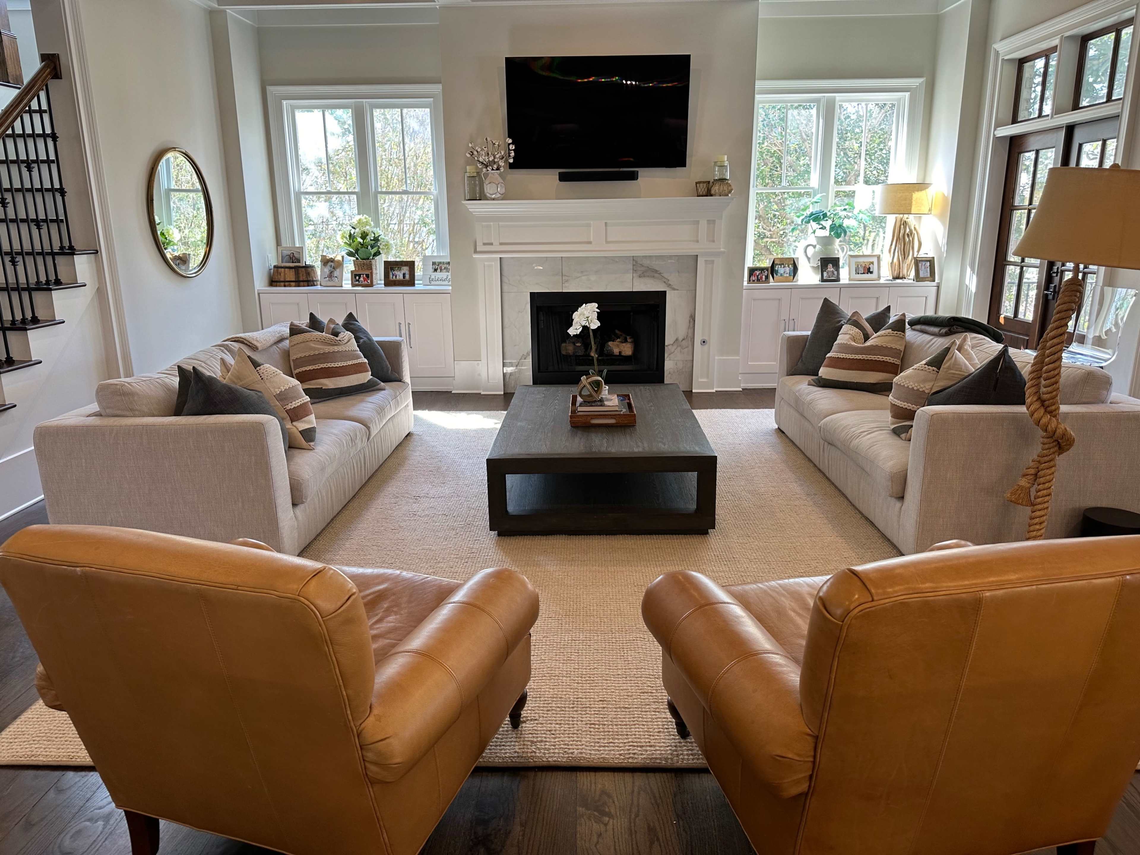 The image shows a living room with two white sofas, two brown leather chairs, and a coffee table on a large area rug, all arranged around a central fireplace and a TV mounted above it.