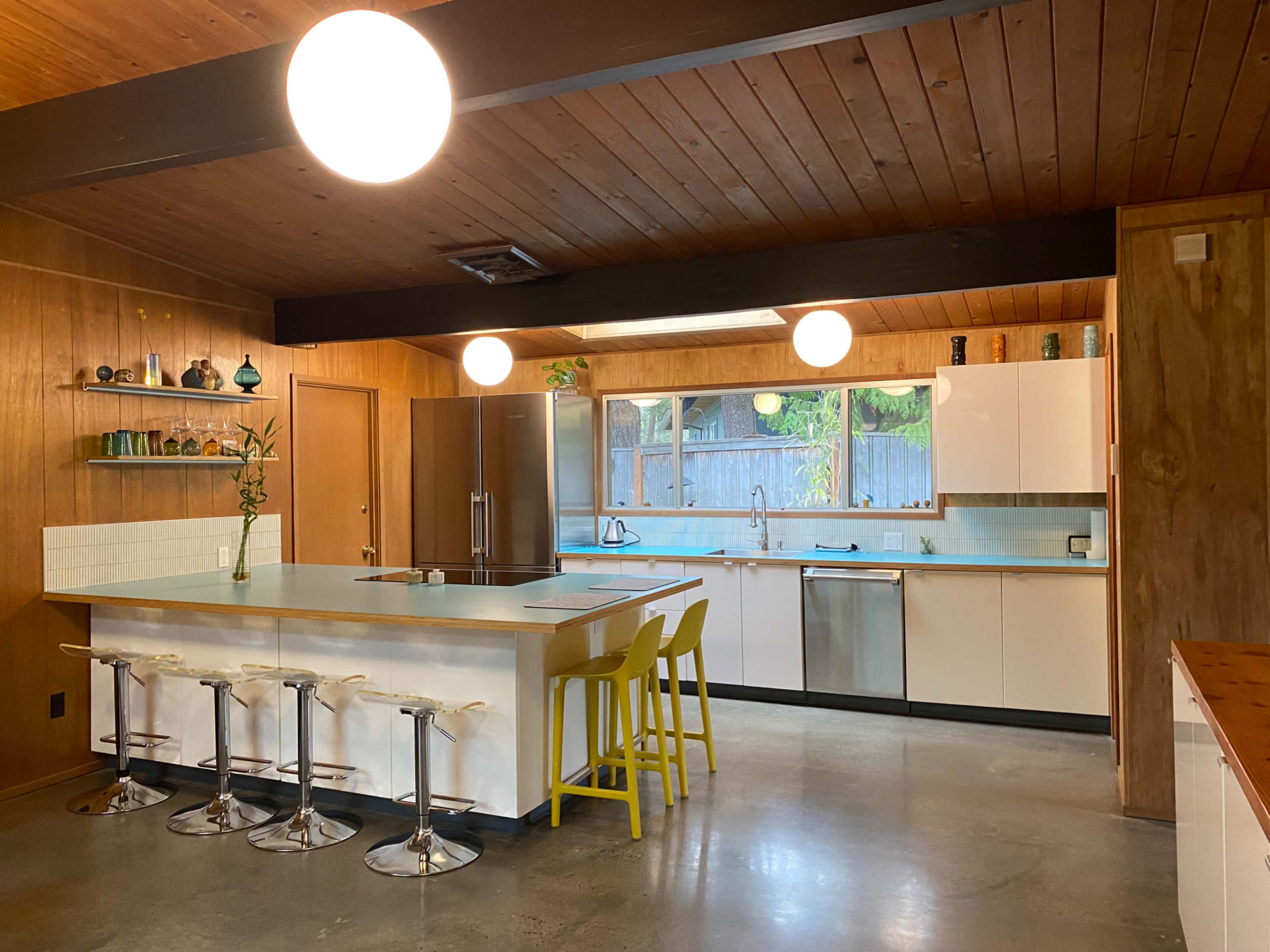 A modern kitchen features a central island with bar stools, sleek cabinetry, and large windows allowing natural light in.