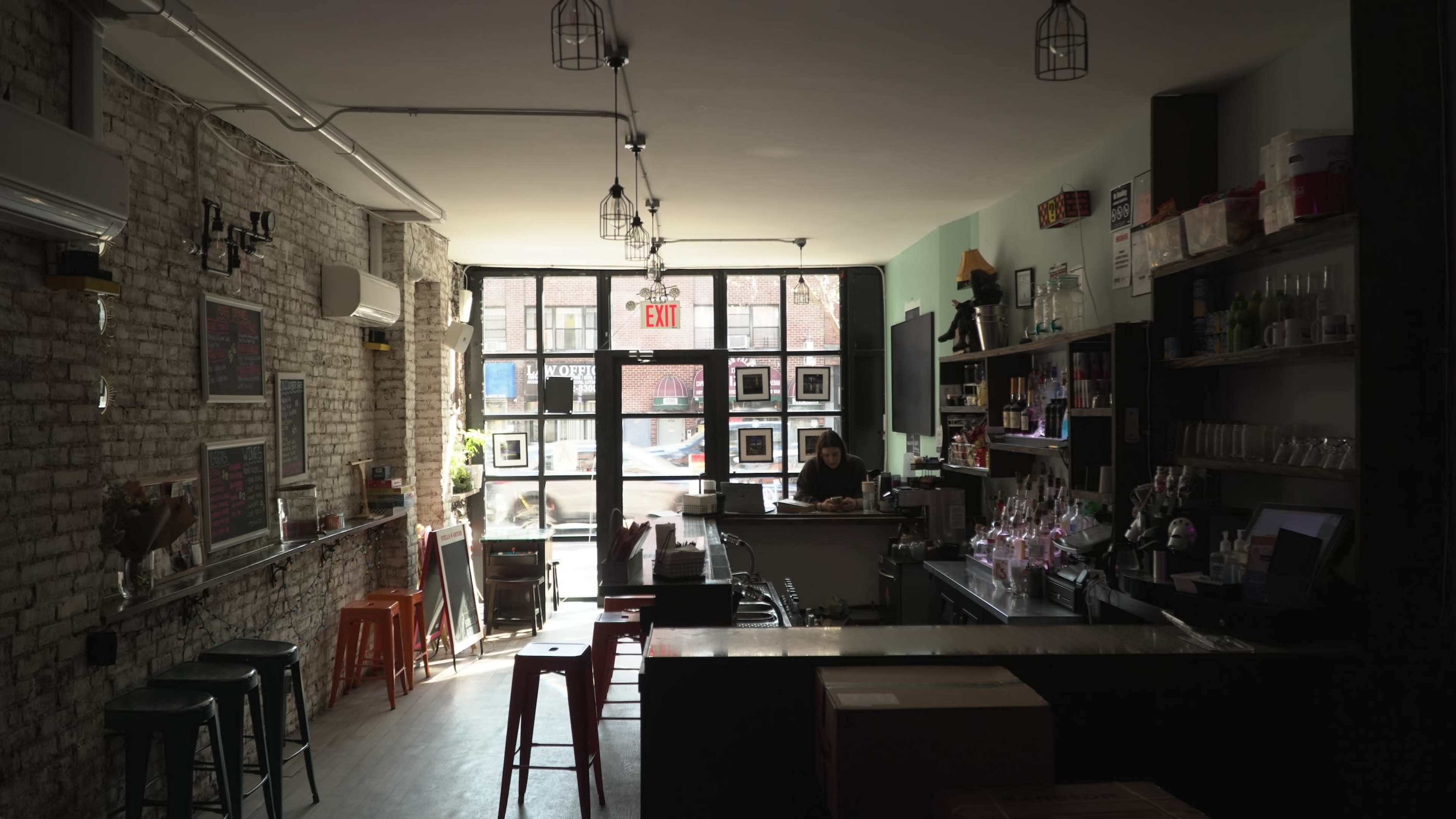 The image shows the interior of a café with a bar area, exposed brick walls, and a large window letting in natural light.