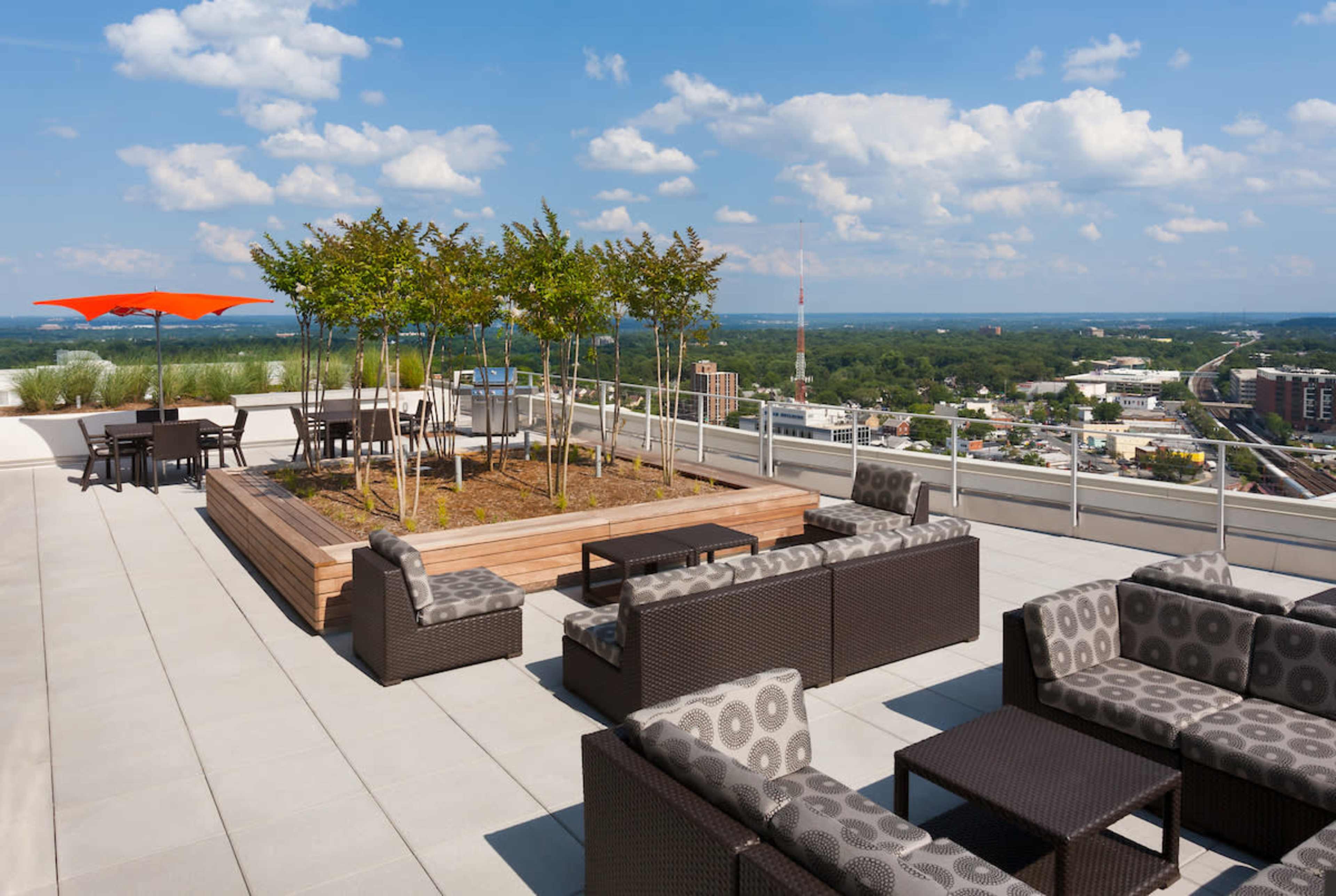 A rooftop terrace featuring modern furniture, a wooden planter with trees, and an orange umbrella, overlooking a cityscape under a partly cloudy sky.