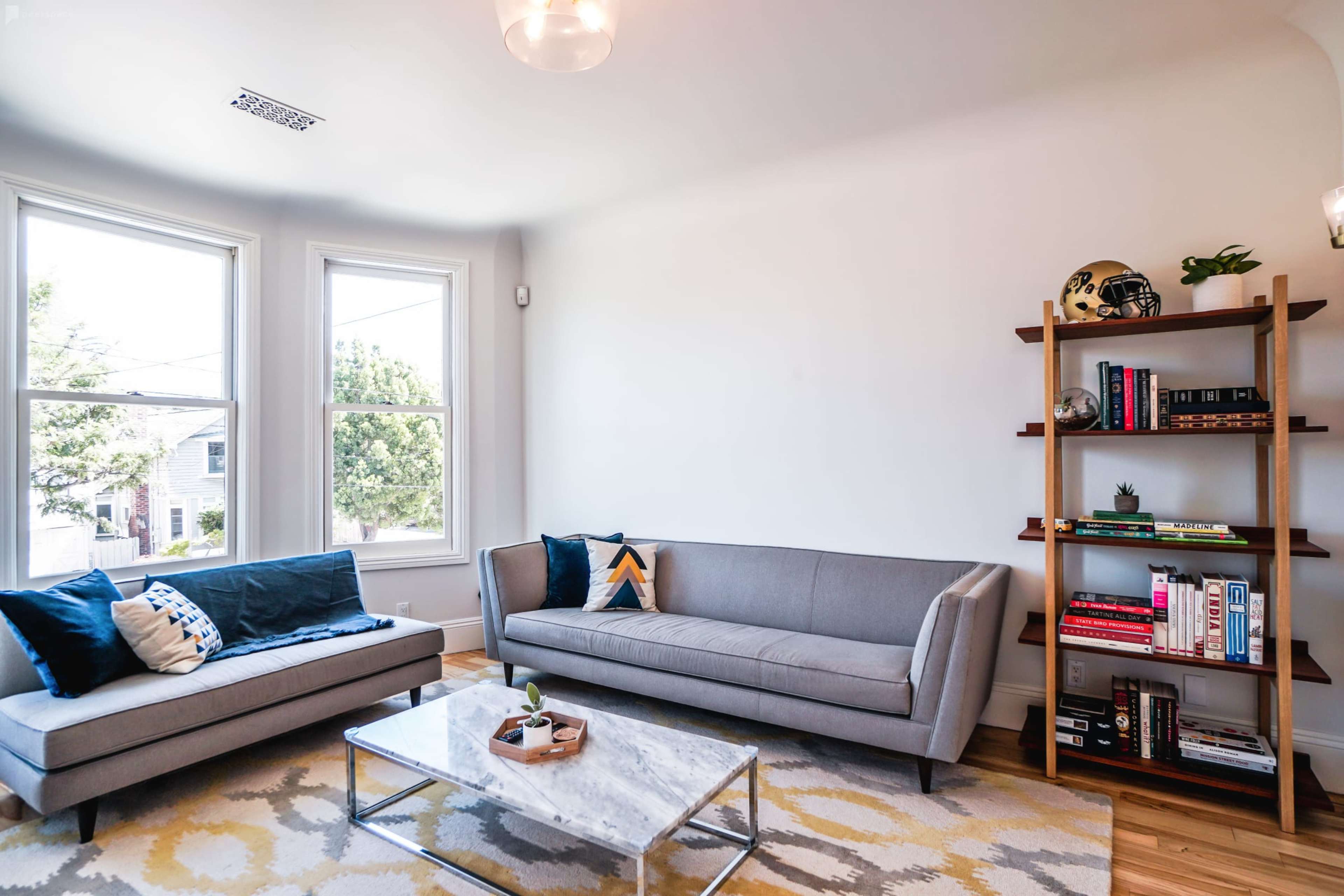 A modern living room features two gray sofas, a marble coffee table, and a bookshelf against a white wall with large windows allowing natural light.