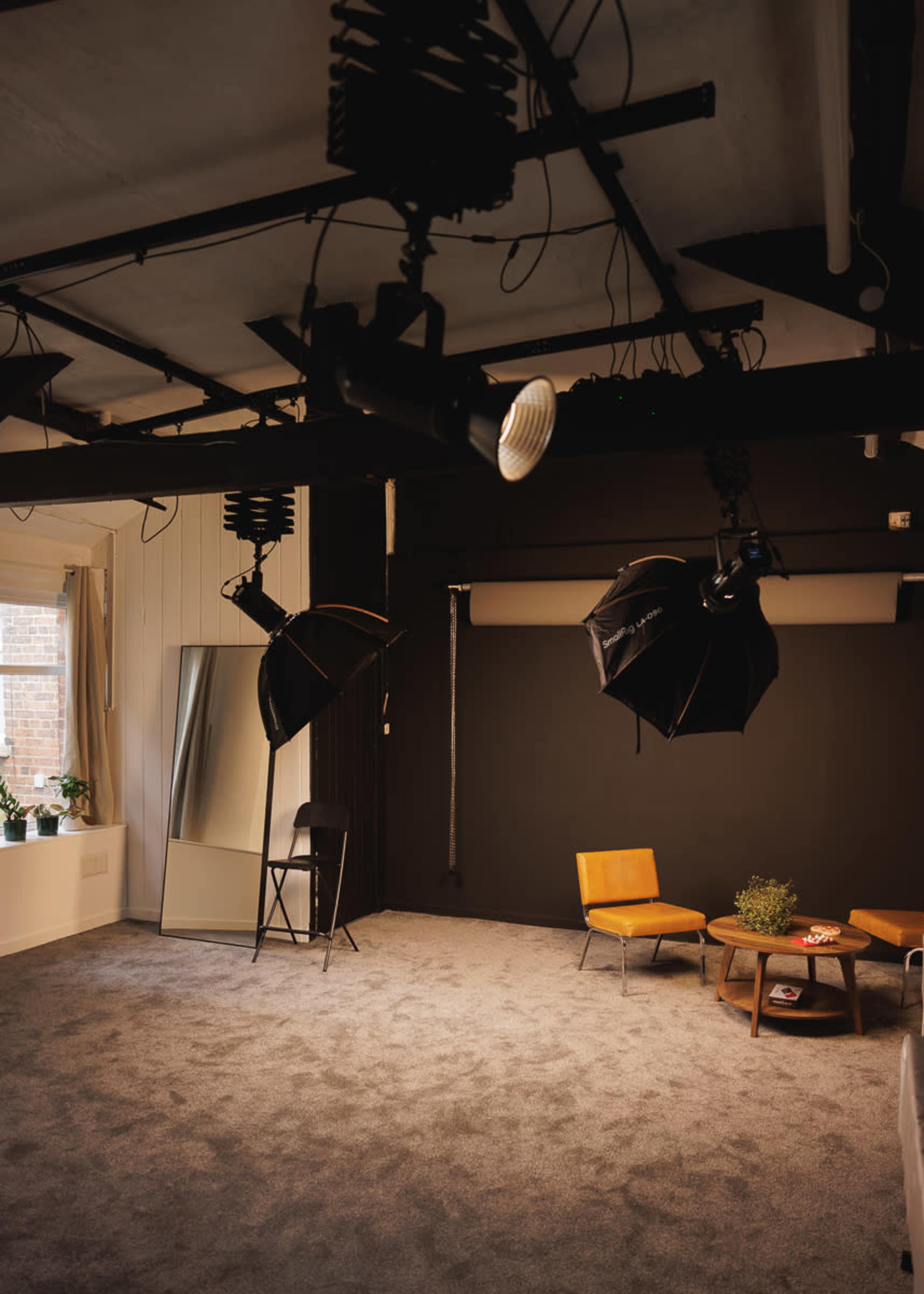 The image shows a spacious photo studio with a dark backdrop, two softbox lights, a mirror, two chairs, and a small table on a carpeted floor.