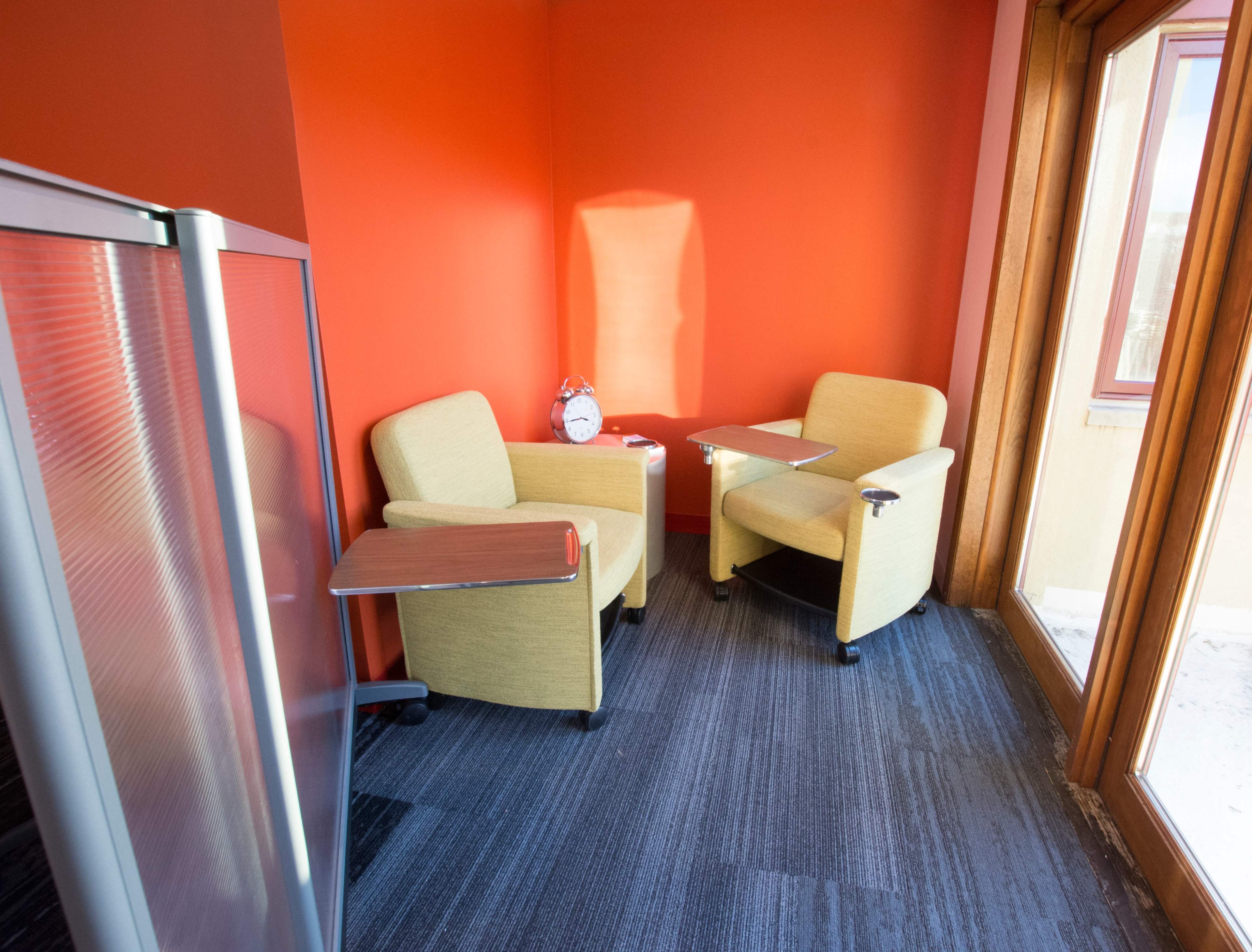 A small seating area with two yellow chairs and a wooden table against a bright orange wall, with a clock on the table and large windows letting in natural light.