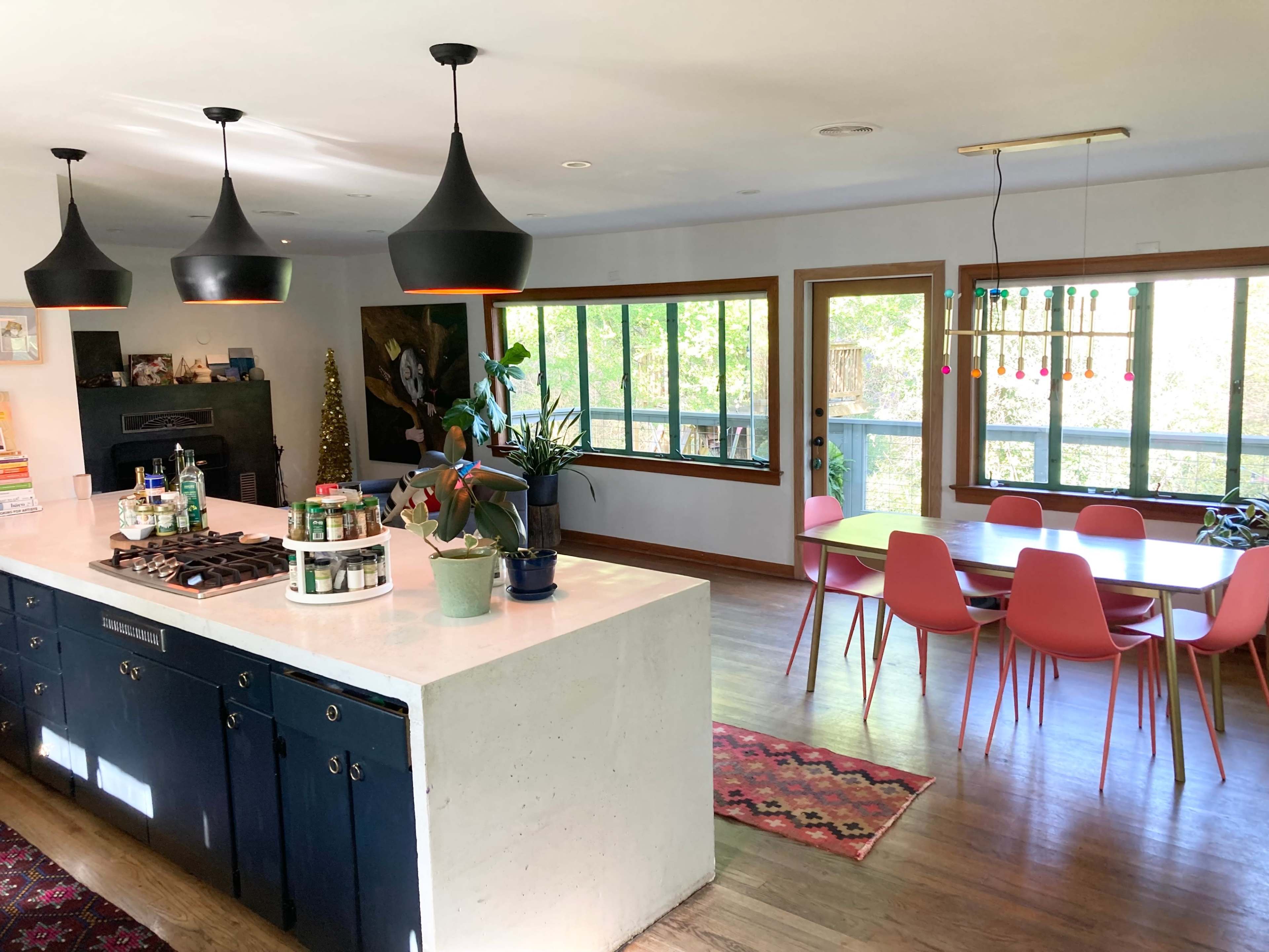 A modern kitchen features a central island with a gas stove, surrounded by plants, leading to a dining area with colorful chairs and large windows that provide natural light.