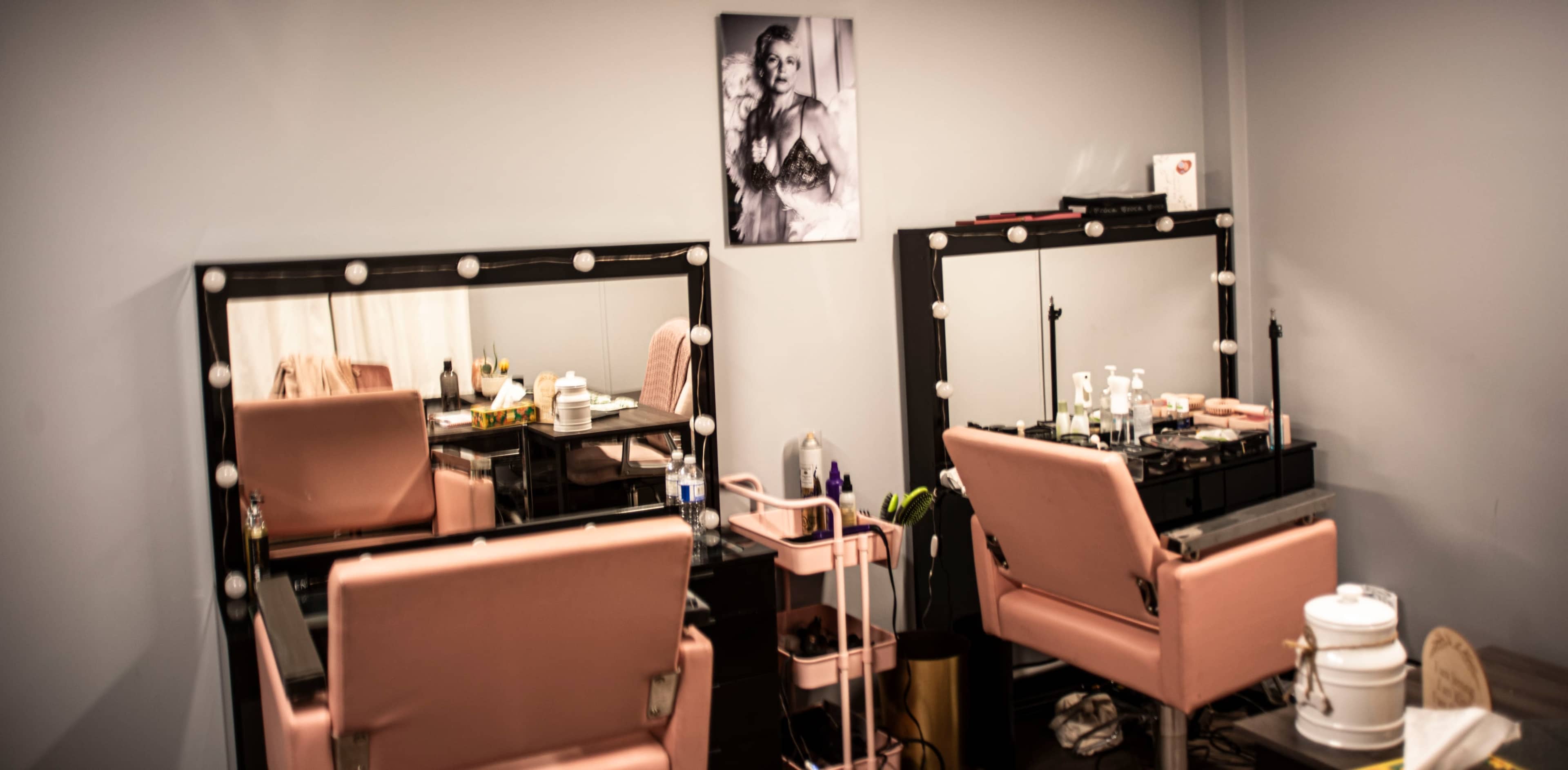 A hair and makeup salon with two pink styling chairs positioned in front of mirrored counters, adorned with makeup tools and a portrait on the wall.