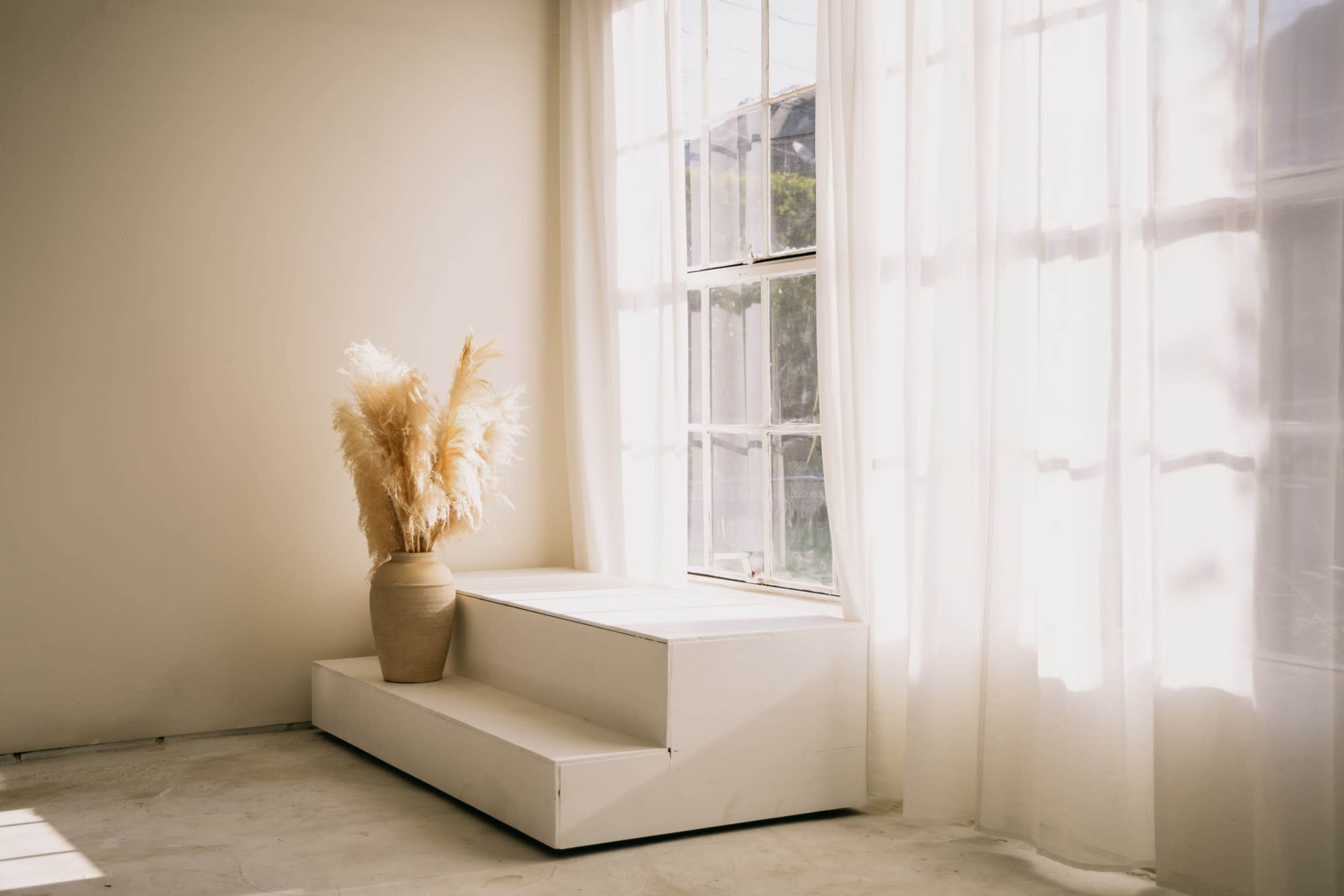 The image shows a minimalist interior with a white staircase and a large vase of dried pampas grass next to sheer curtains.