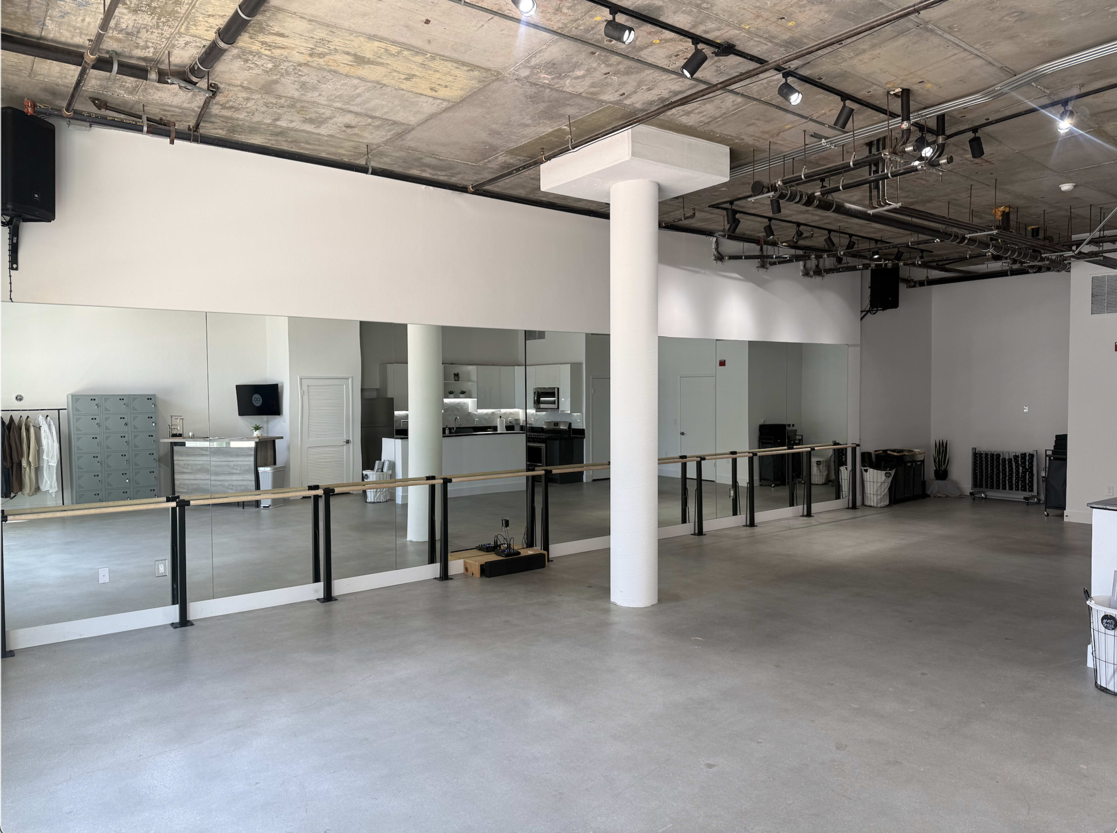 The image shows a spacious, minimalist studio with large mirrors lining one wall and various furnishings, including a counter and storage units, visible in the background.