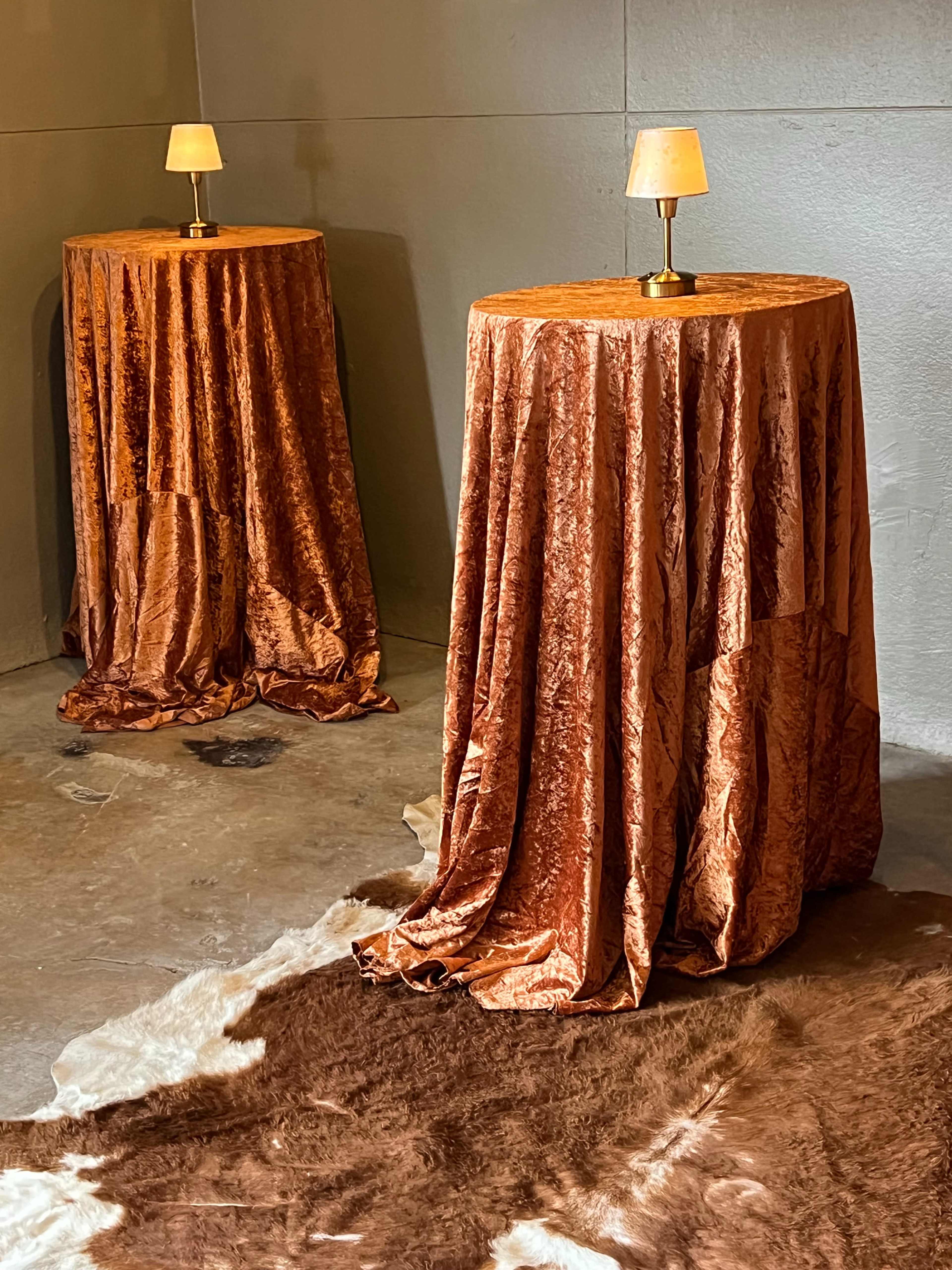 Two circular tables draped with orange velvet cloths stand on a textured floor, each topped with a small lamp.