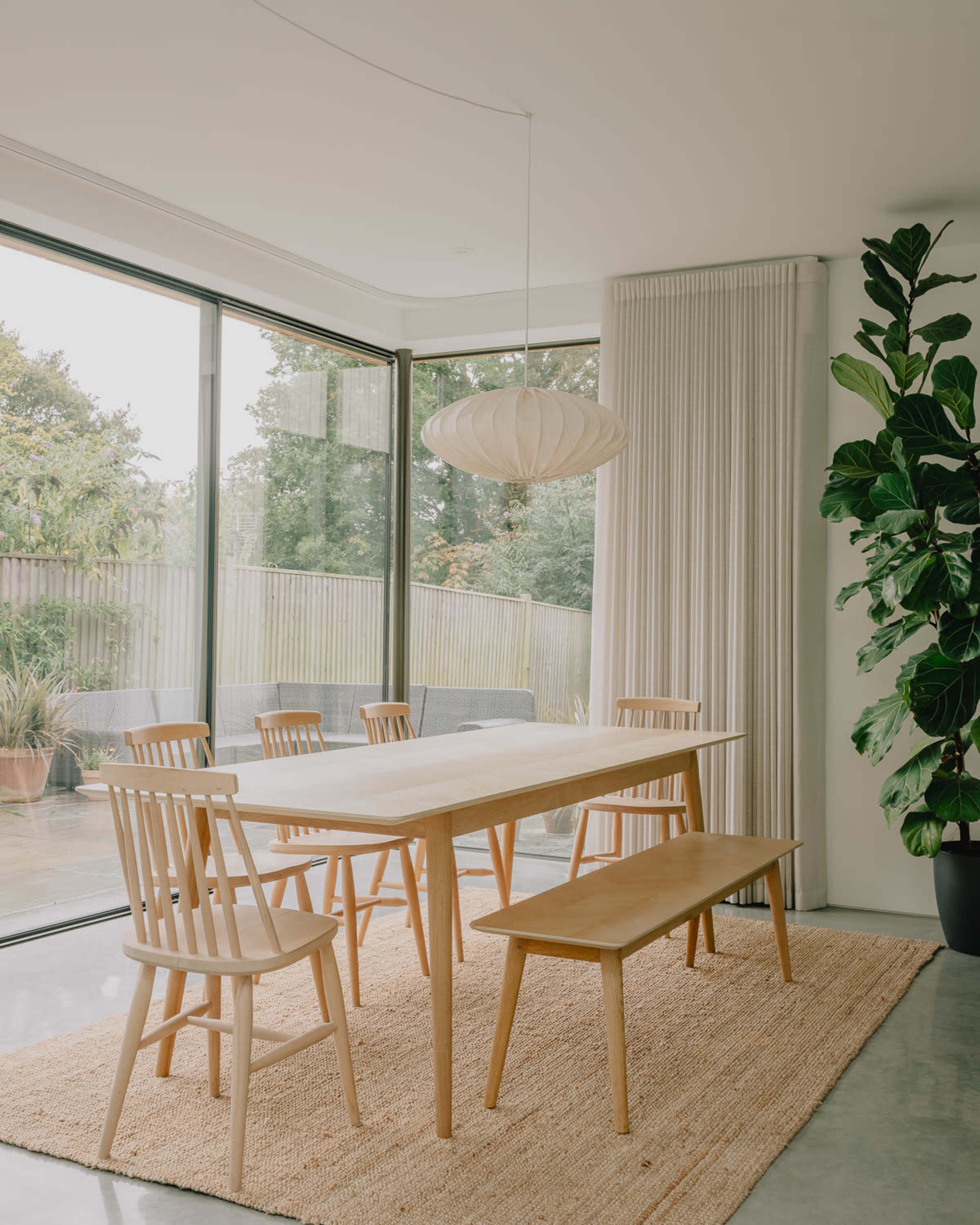 A modern dining area features a long table surrounded by chairs and a bench, with large windows that overlook a green outdoor space.