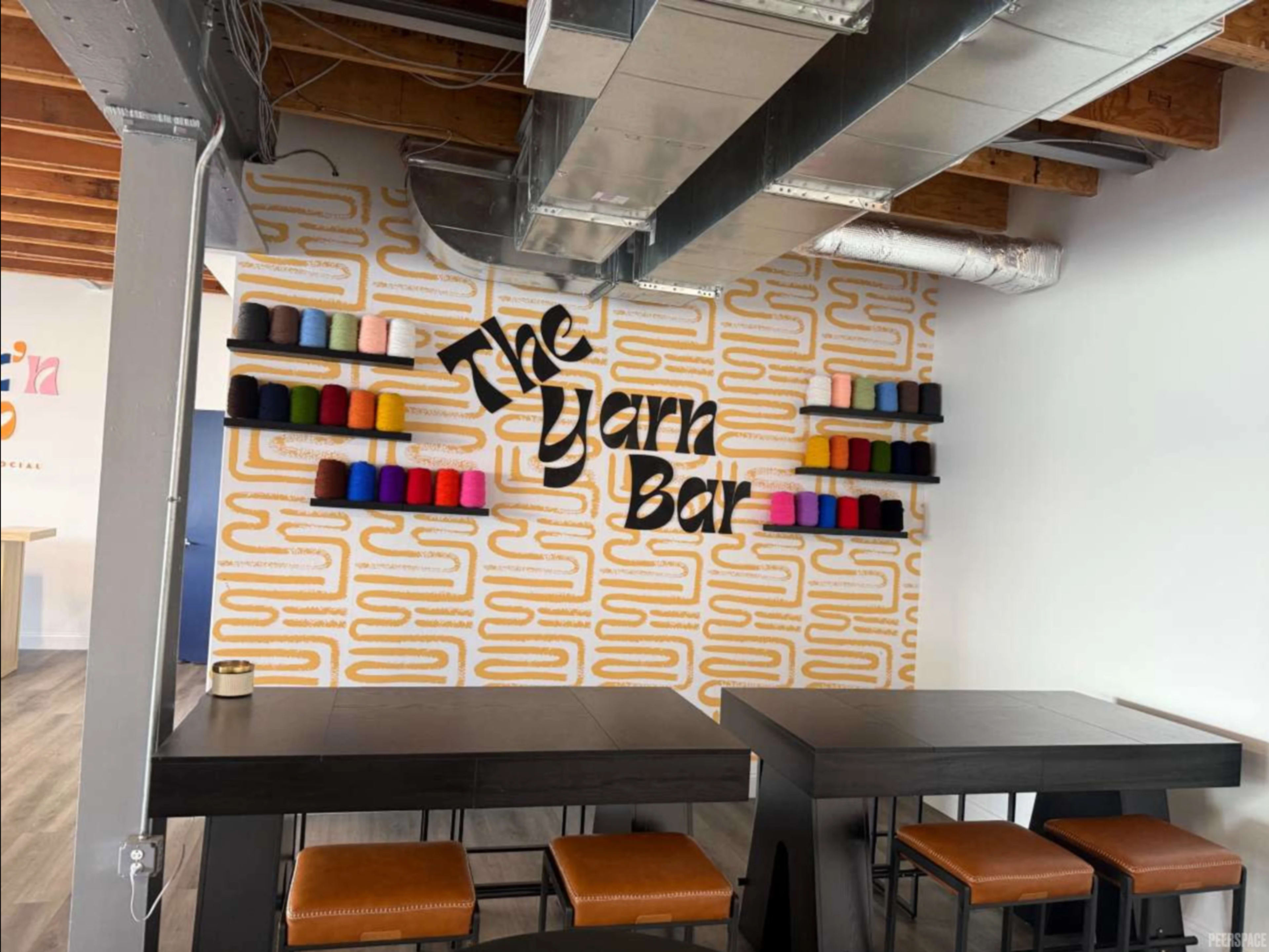 The image shows a modern interior of a space called "The Yarn Bar," featuring a wall with colorful yarn spools arranged on shelves and two black tables with orange seating.