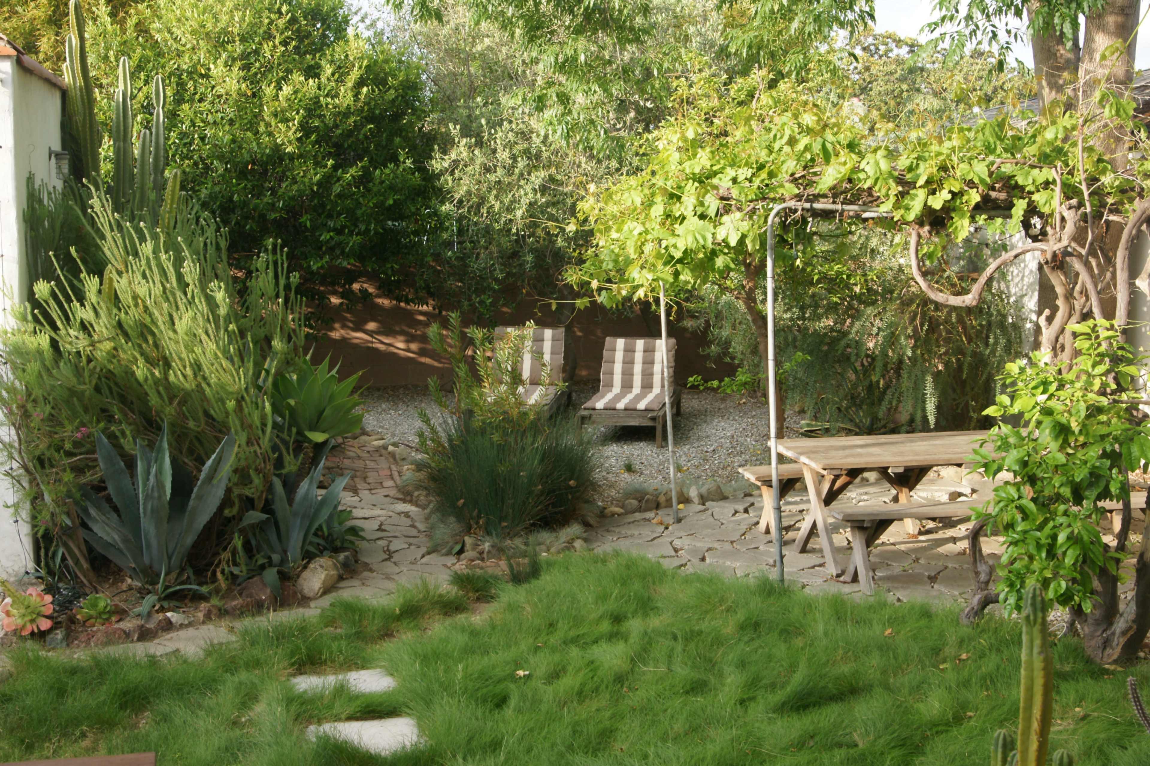 The image depicts a tranquil garden with lounge chairs, a wooden table, various plants, and a stone pathway.