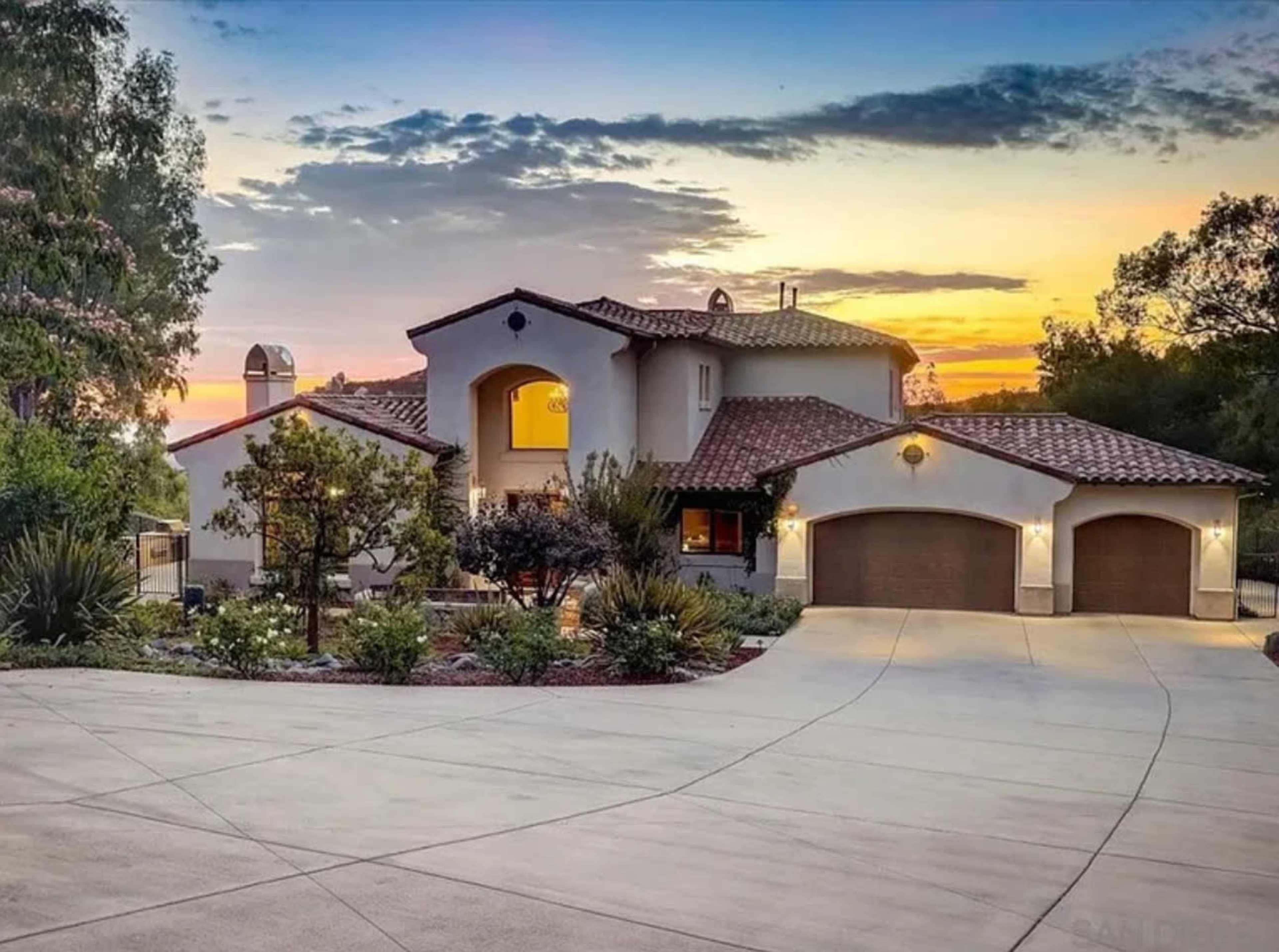 The image shows a large, two-story house with a red-tiled roof, flanked by trees and shrubs, set against a colorful sunset.