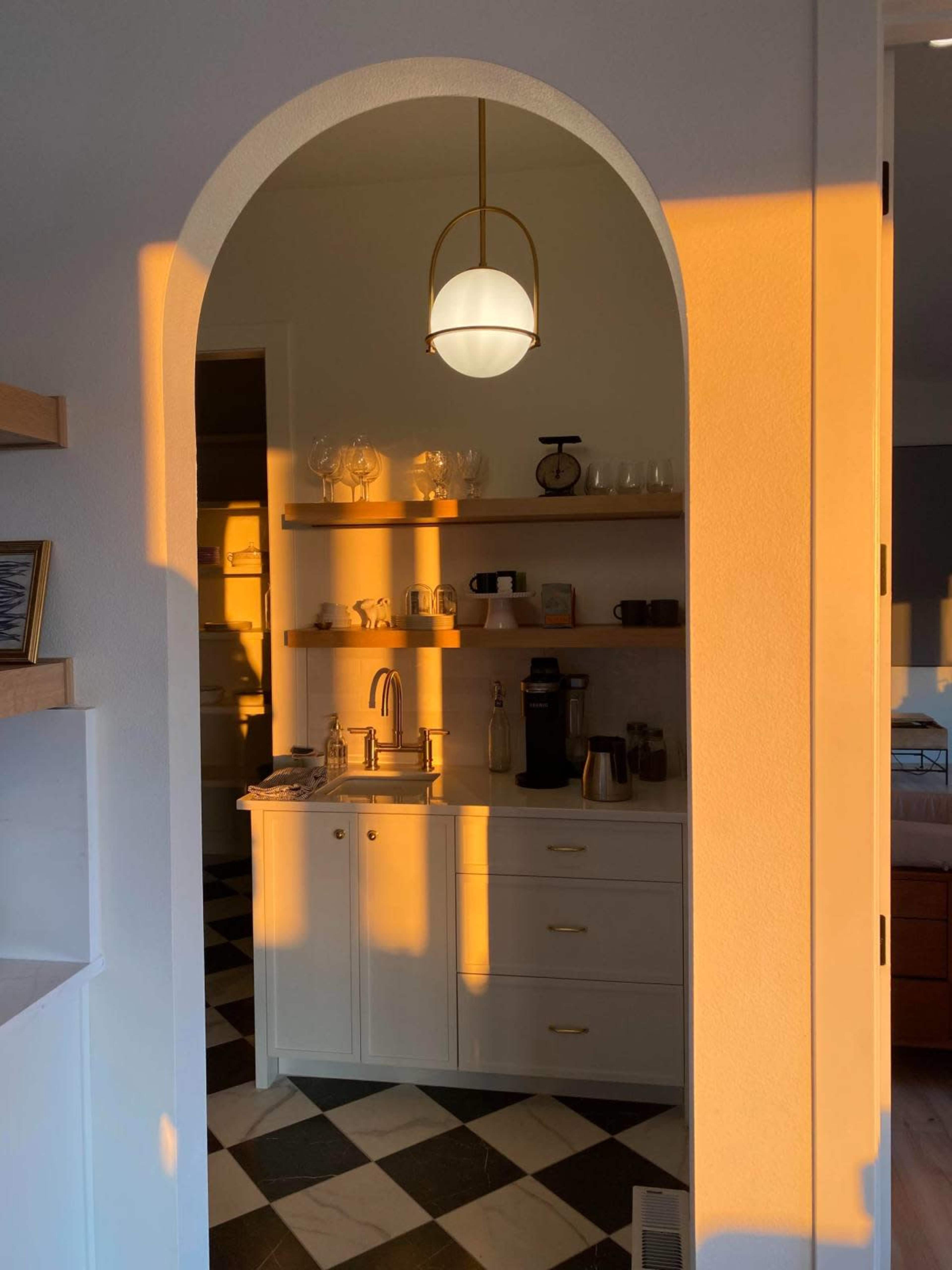 A warm glow from the setting sun casts shadows on a small kitchen area featuring a sink and shelves filled with glassware.