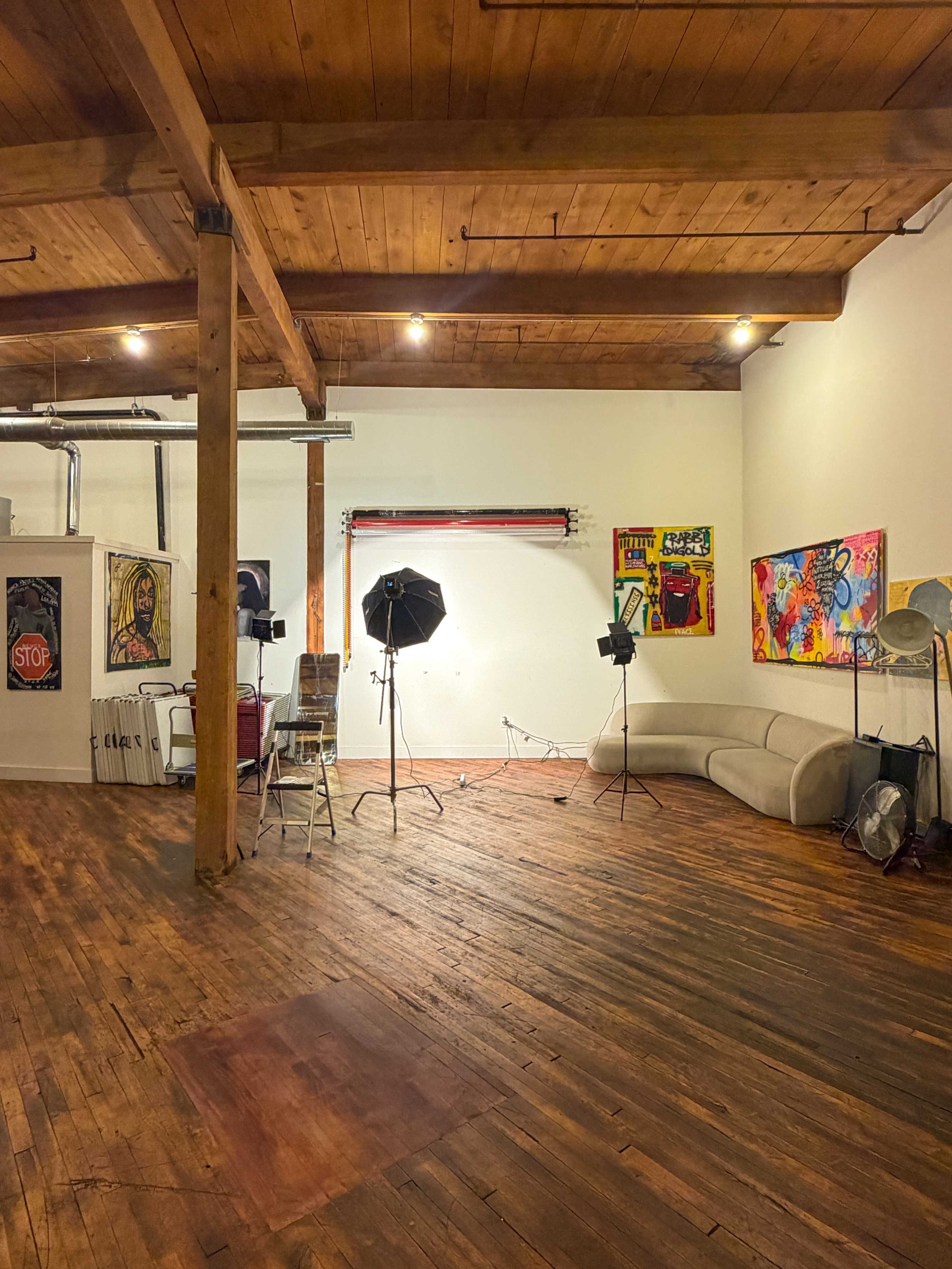 The image shows a spacious room featuring wooden floors, high ceilings, a curved couch, and several photography lights set up for a shoot.