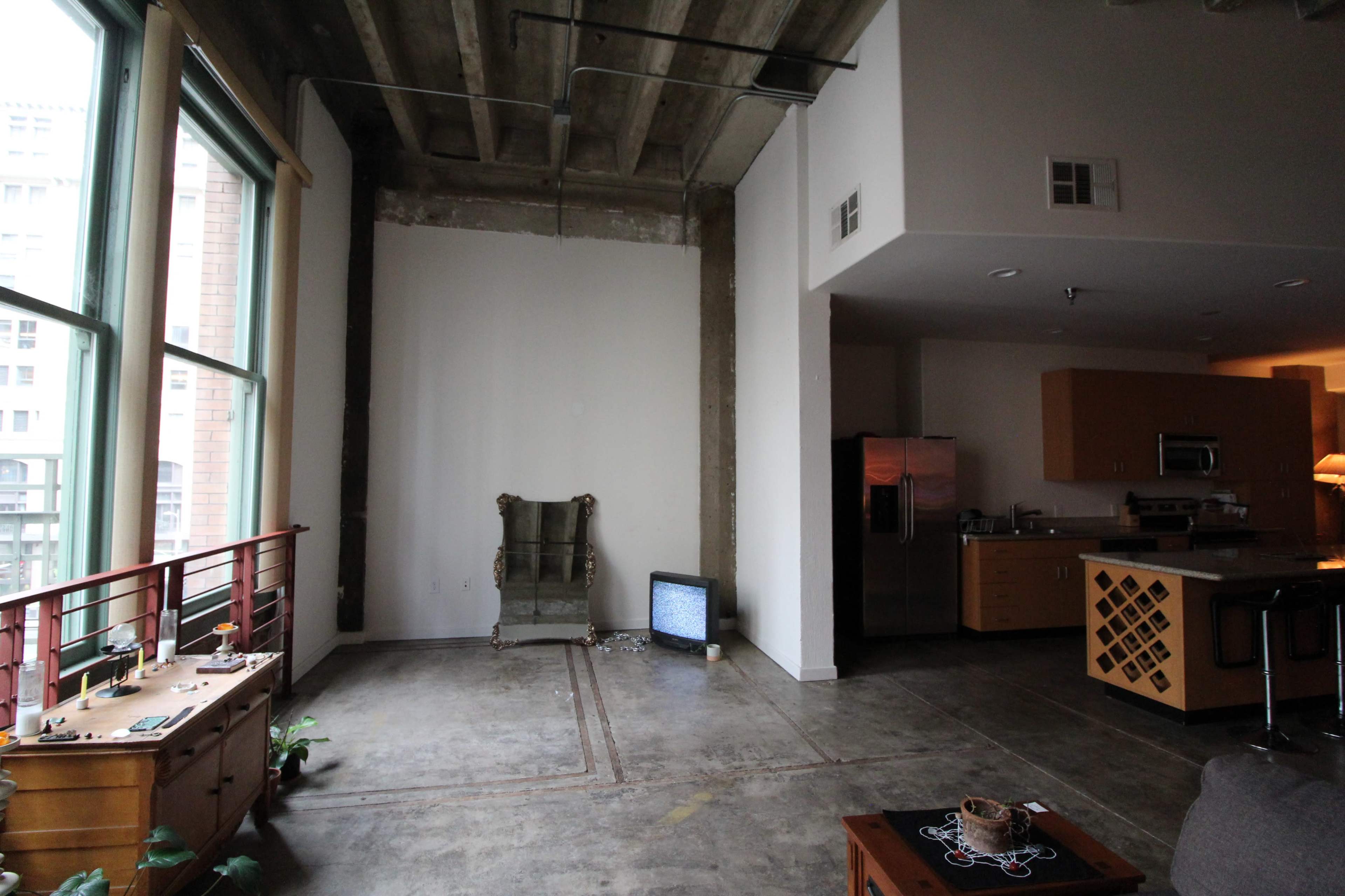 The image shows a spacious industrial-style living area with a green chair, a vintage television set, and a modern kitchen in the background.