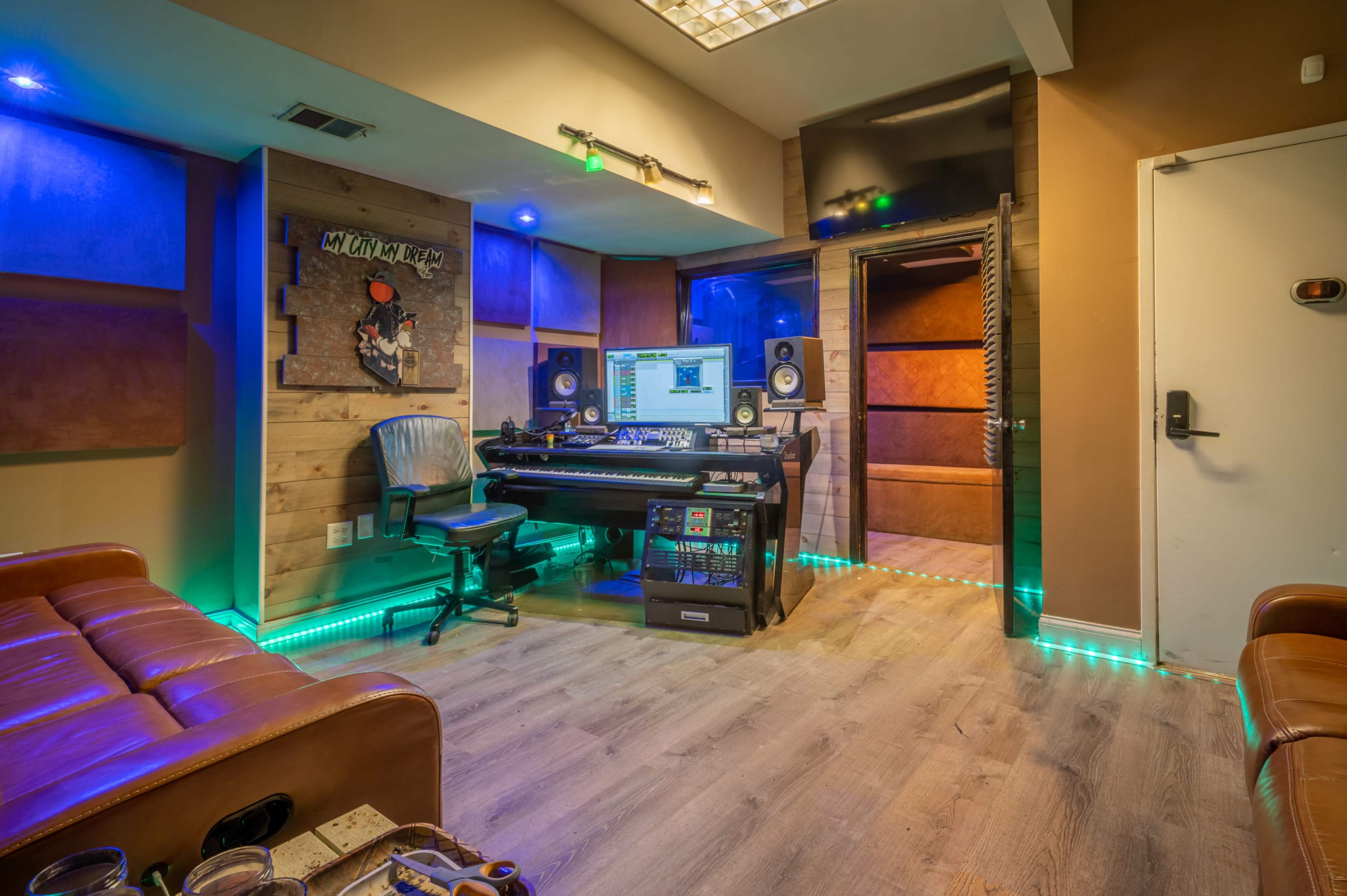 The image shows a small music recording studio featuring a computer, mixing desk, and seating area with ambient lighting.