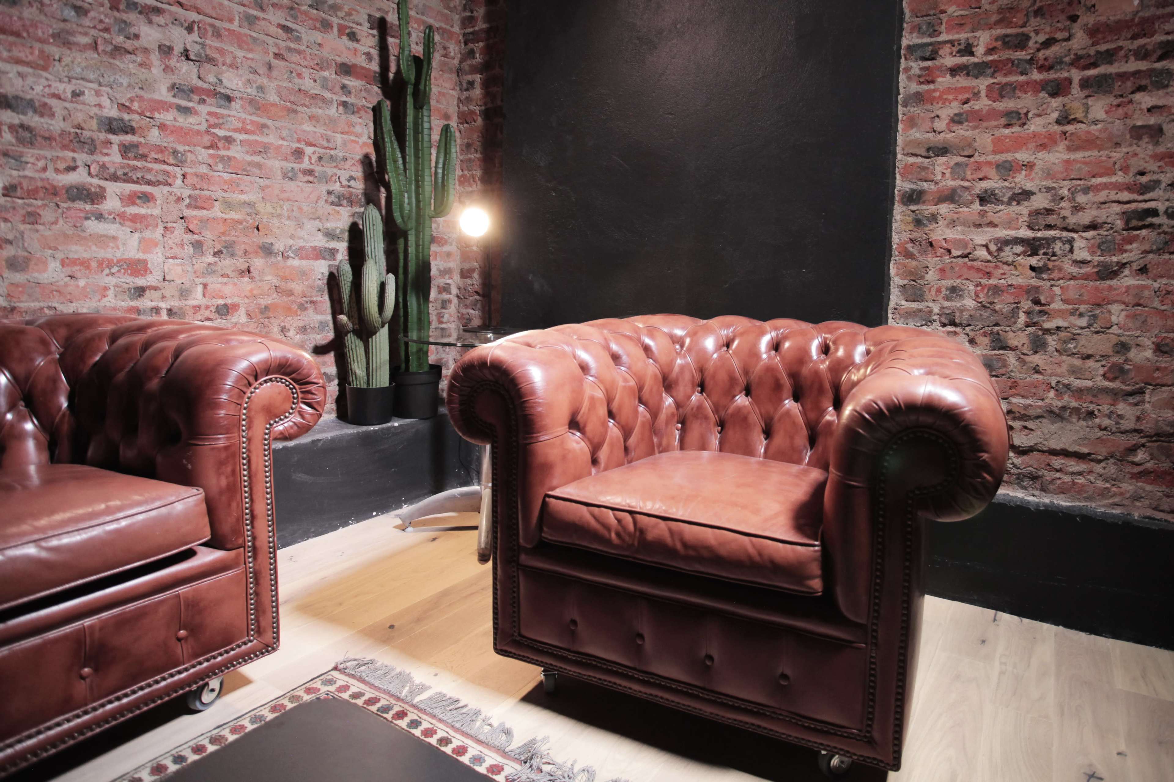 The image shows two brown leather tufted armchairs positioned near a potted cactus in a modern, industrial-style room with exposed brick walls.