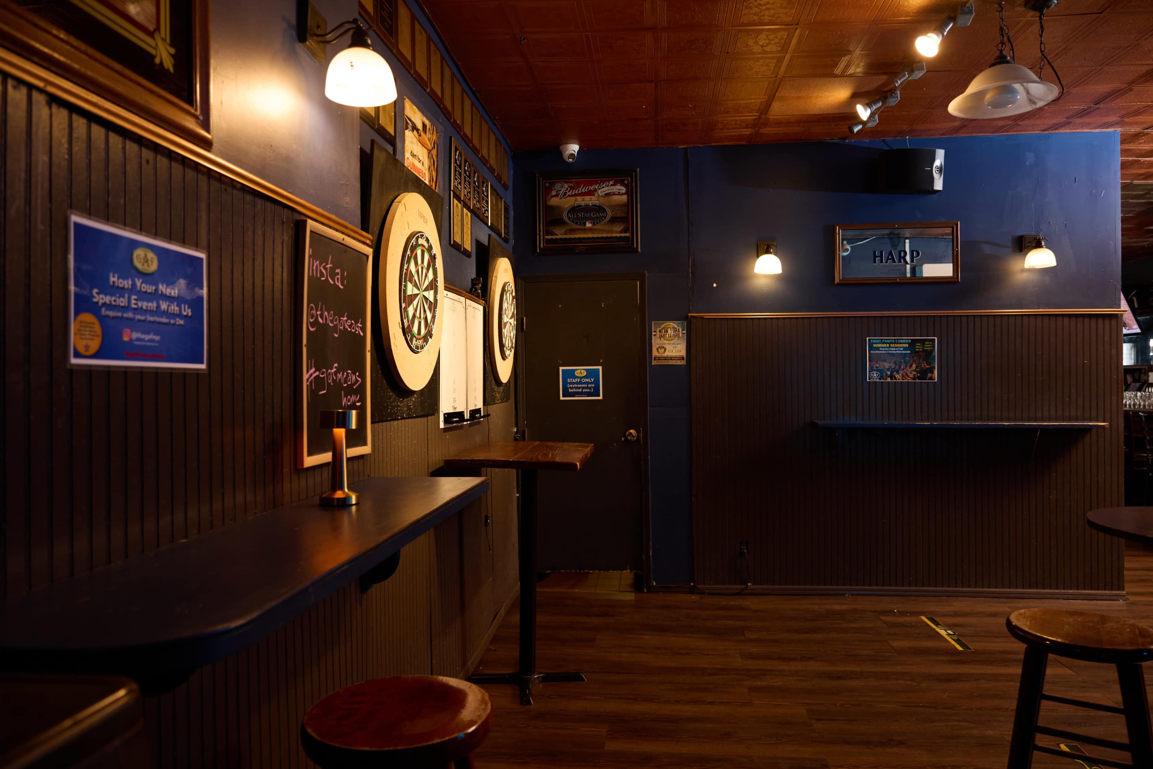 The image shows a dimly lit bar interior featuring dartboards and a simple seating area with high tables and stools.