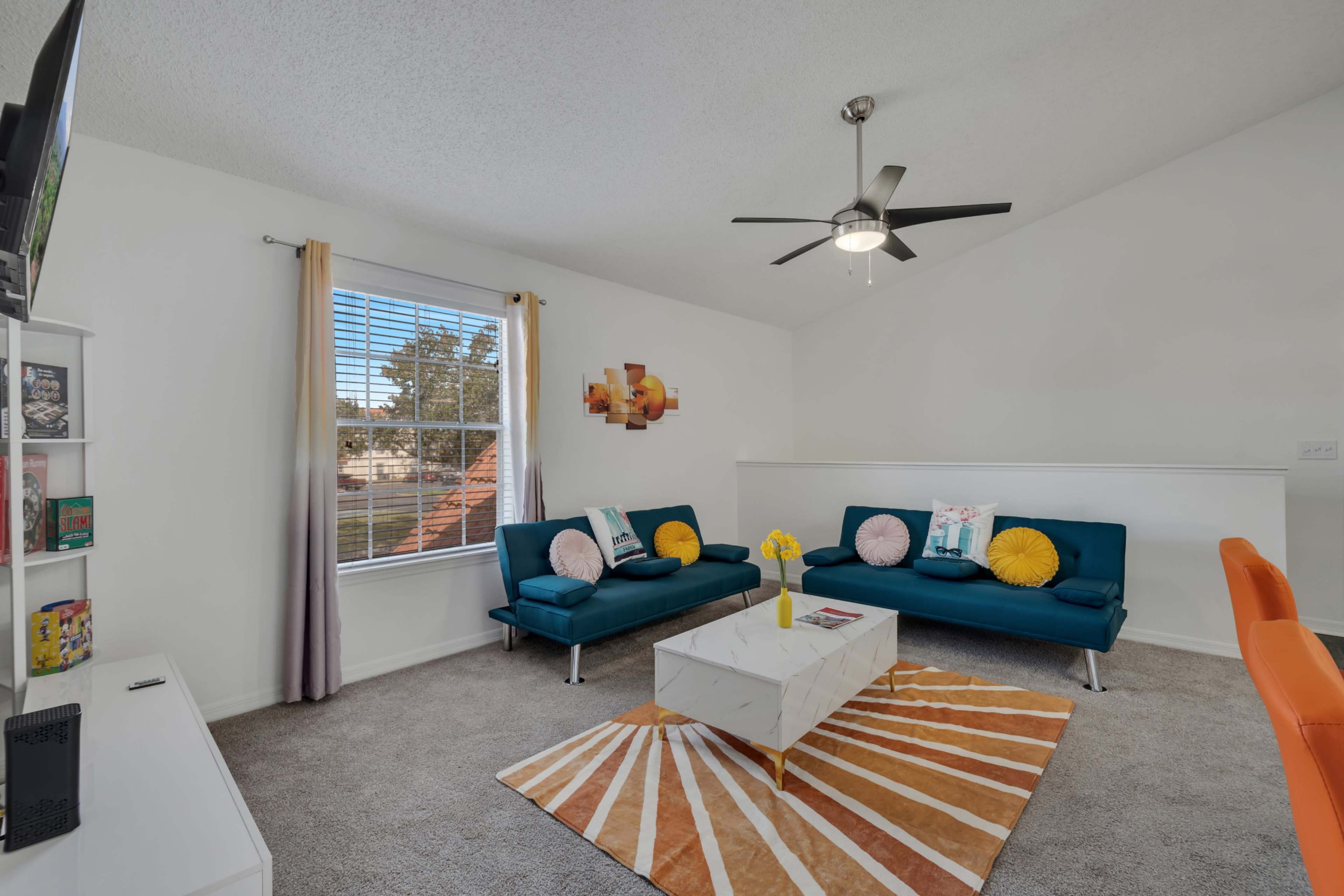 The living room features two teal sofas, a white coffee table, a patterned rug, and a ceiling fan, with a large window allowing natural light to illuminate the space.