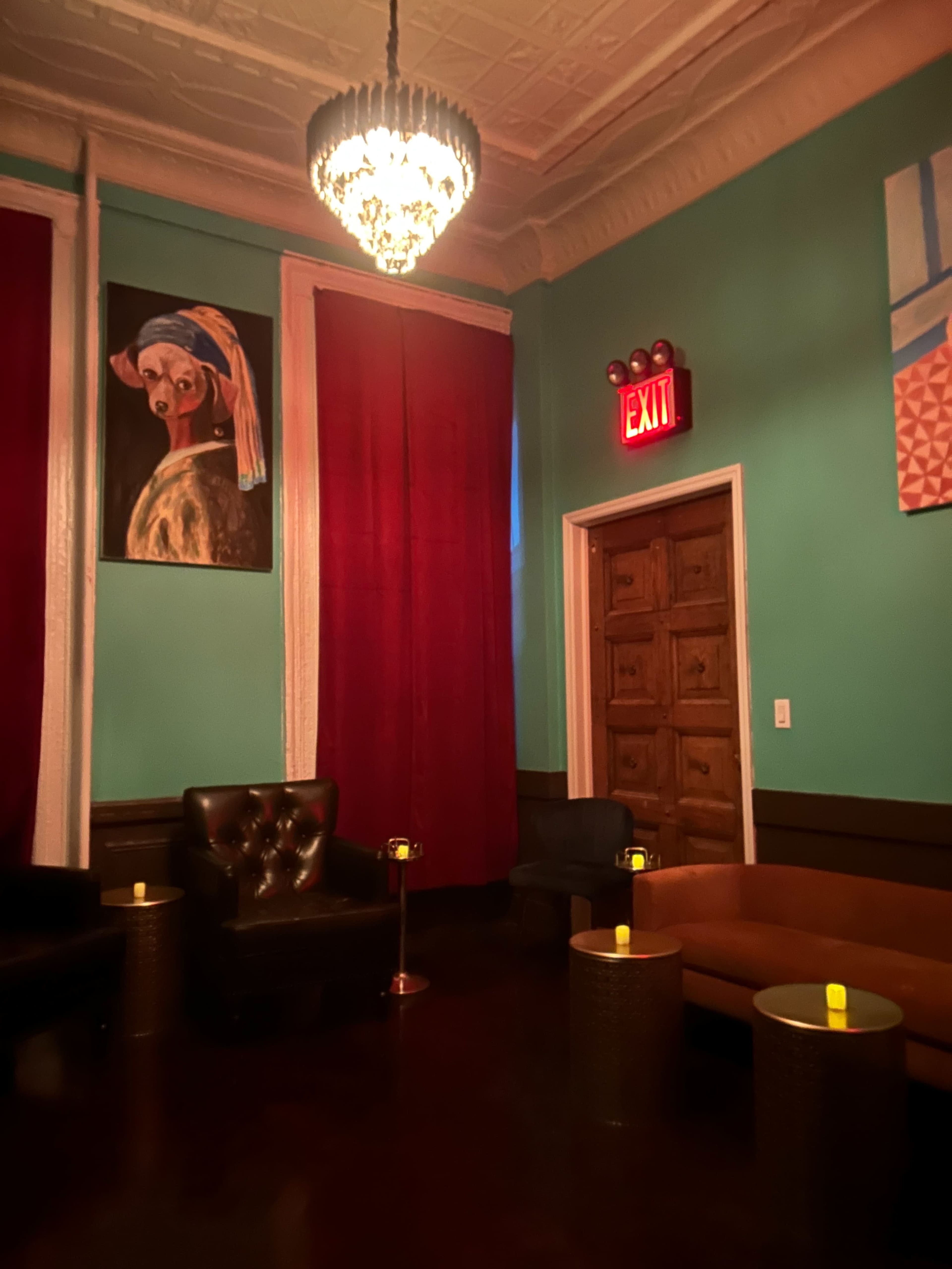 The image shows a cozy lounge area with dark furniture, a large painting on the wall, red curtains, and a chandelier, creating an intimate atmosphere.