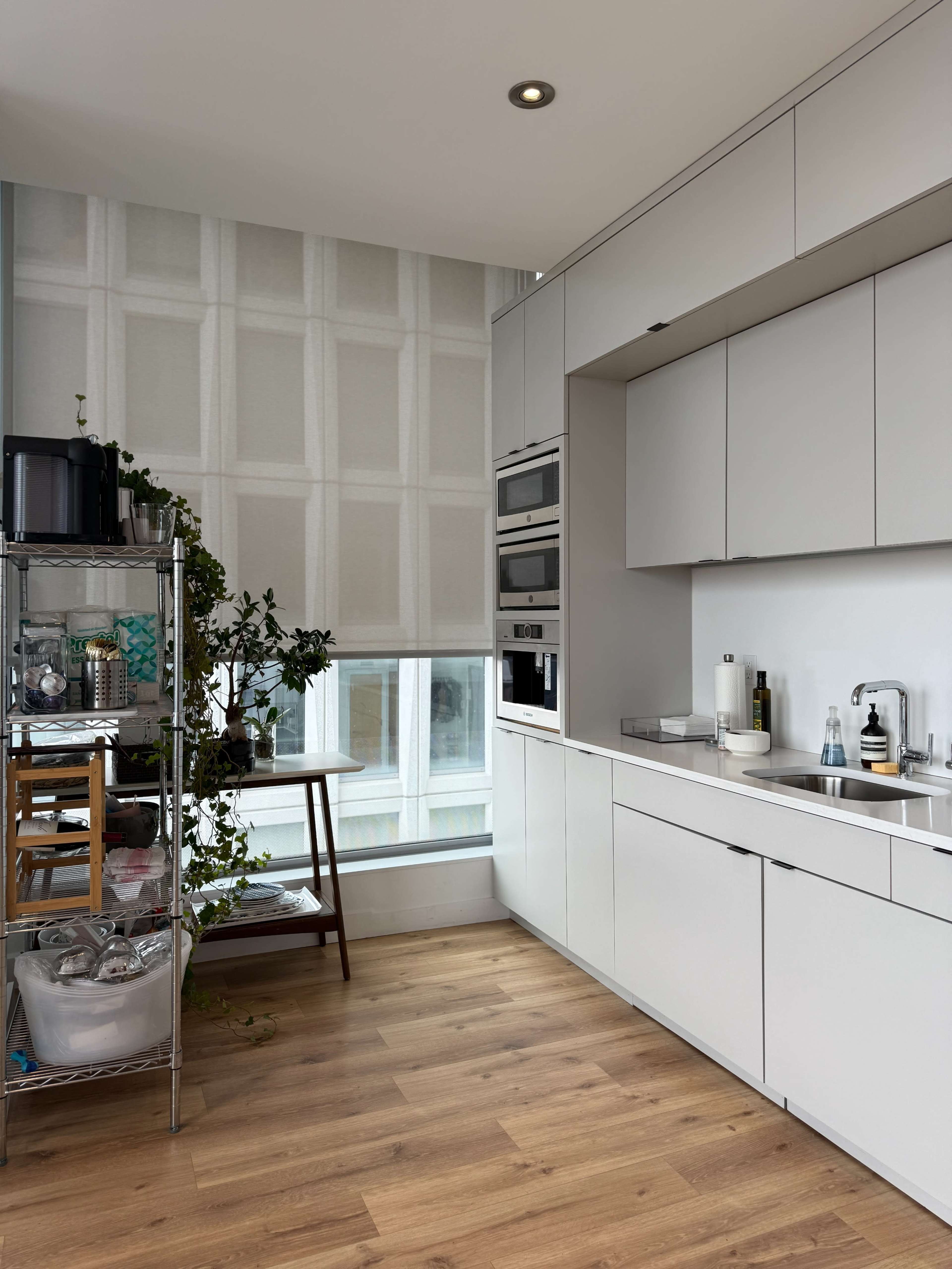 The image shows a modern kitchen with sleek white cabinetry, stainless steel appliances, and a small wooden table by a large window, framed by plants.