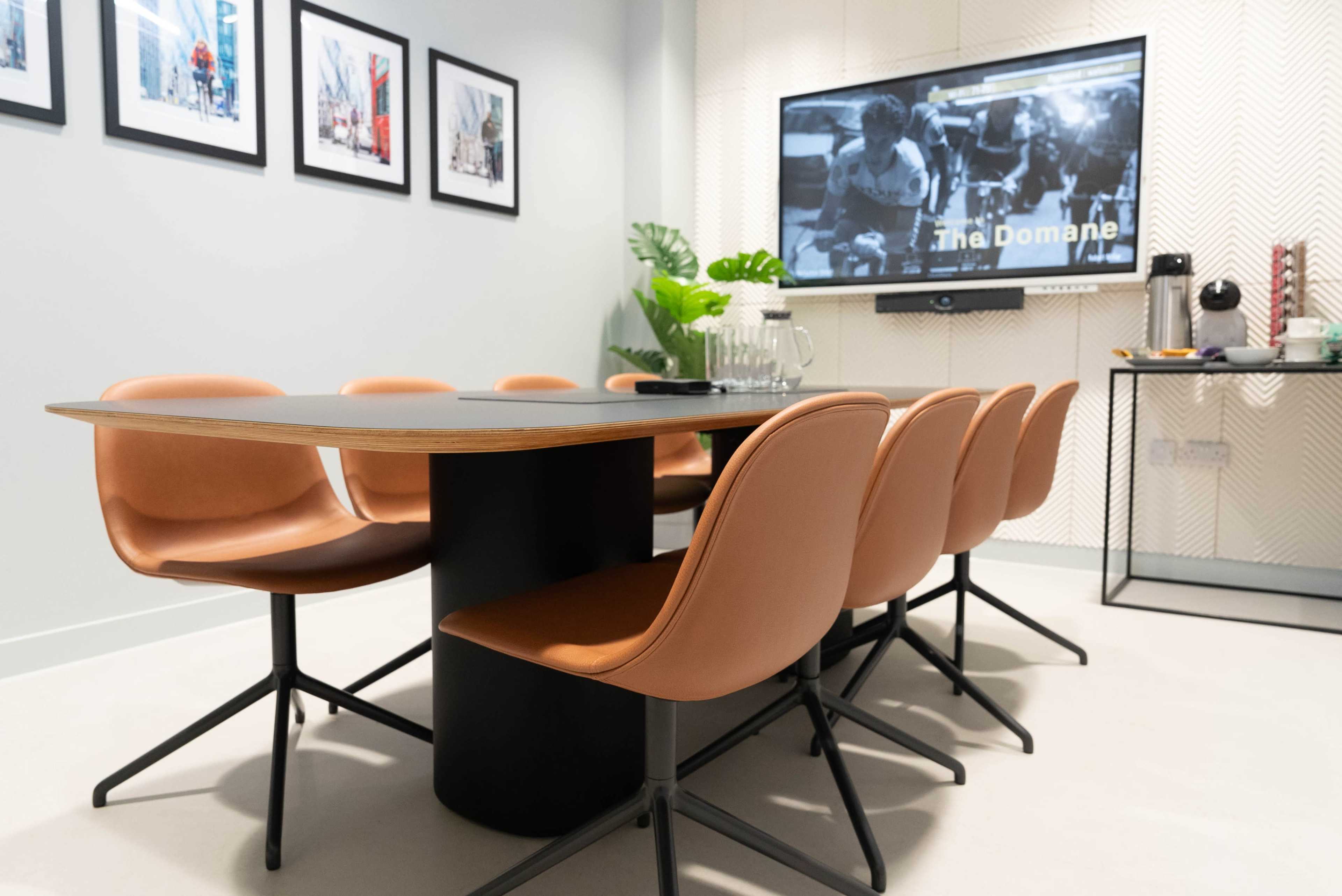 A modern conference room features a large table surrounded by six brown chairs, with a screen displaying a black-and-white image on the wall.